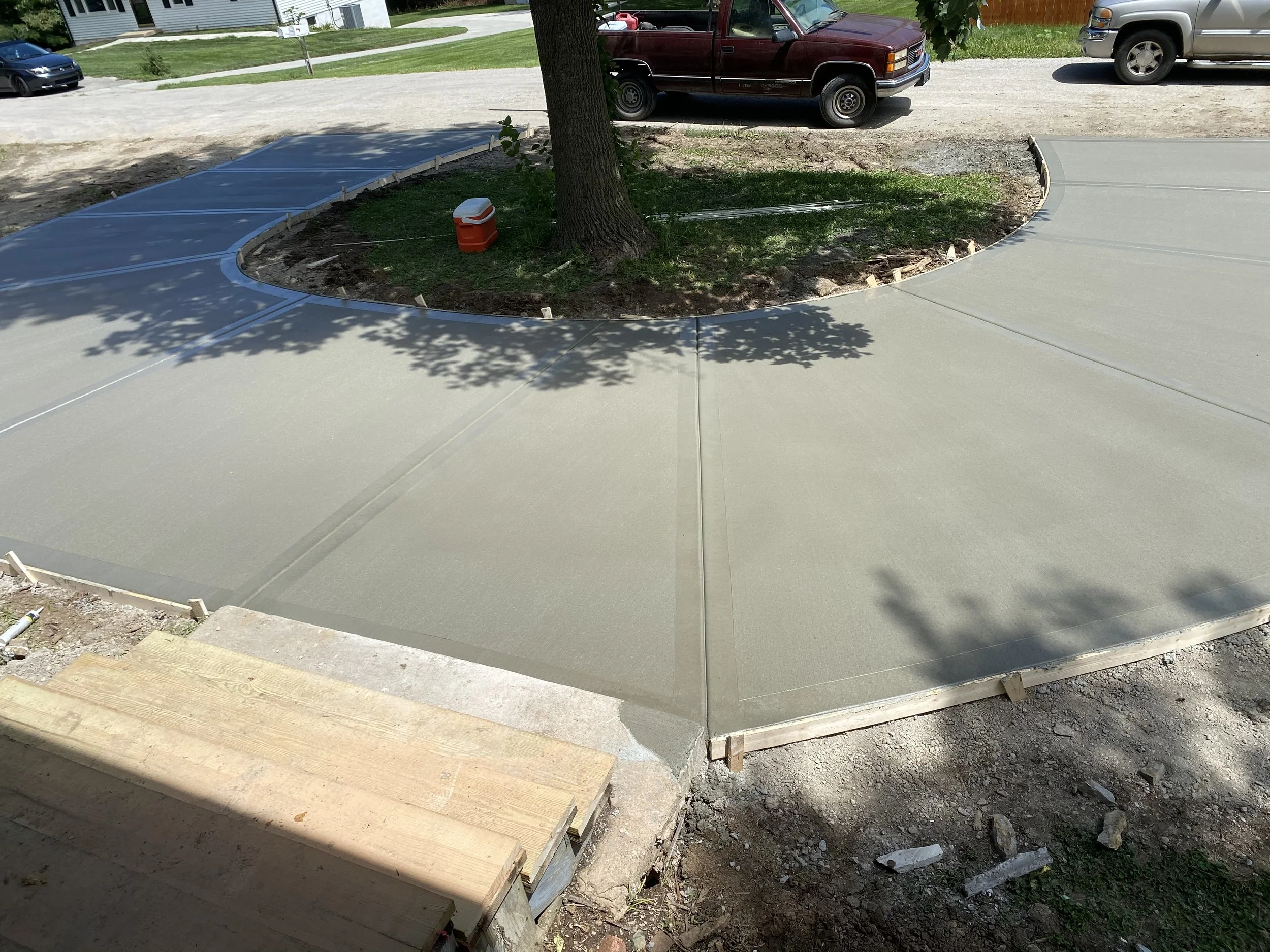 Freshly poured concrete sidewalk curving around a tree in a residential yard. Construction materials and tools are visible, with parked cars in the background.