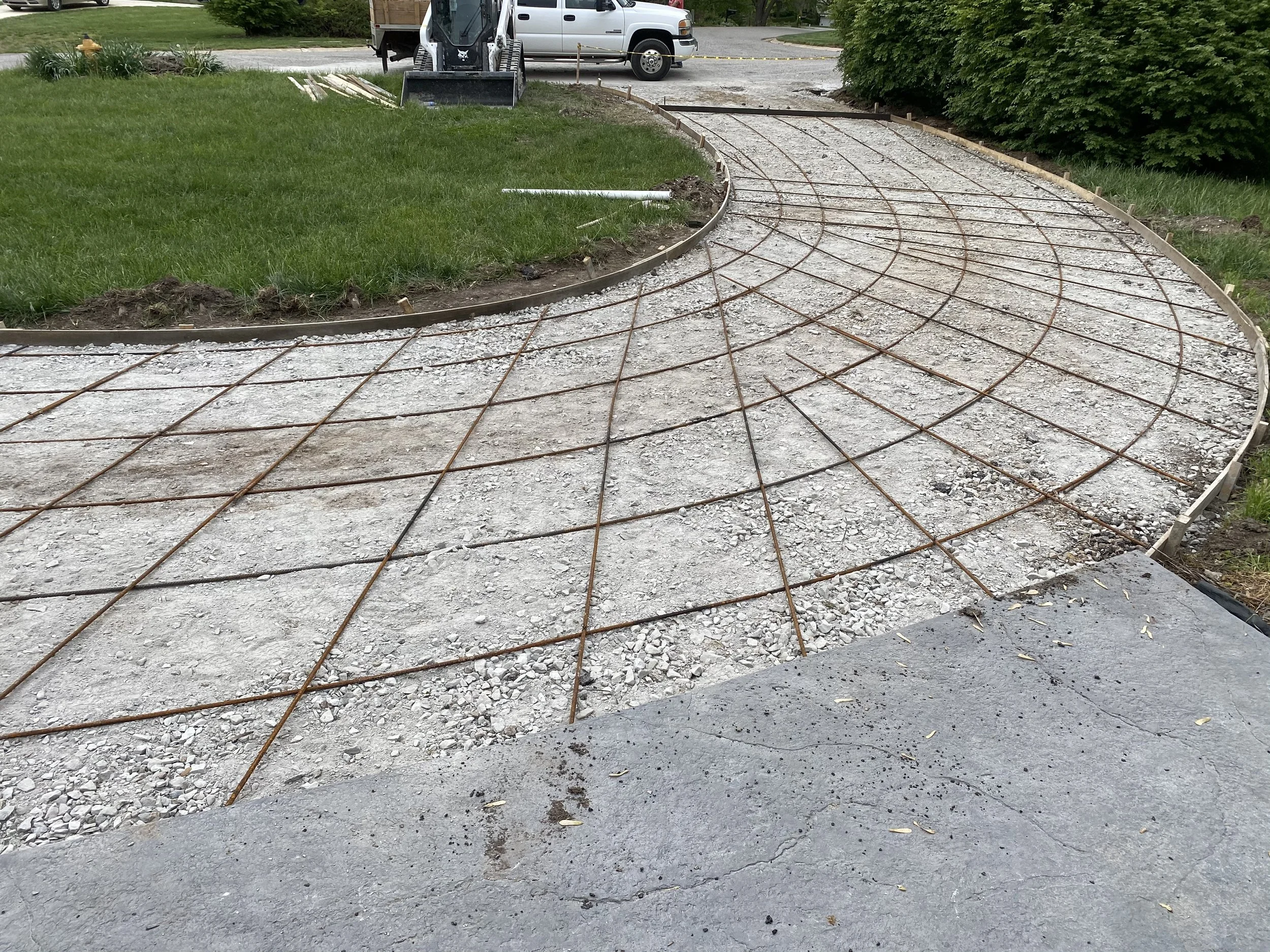 A curved pathway under construction with gravel and metal rebar, bordered by a wooden frame, with grass and bushes on either side, and construction vehicles and equipment in the background.