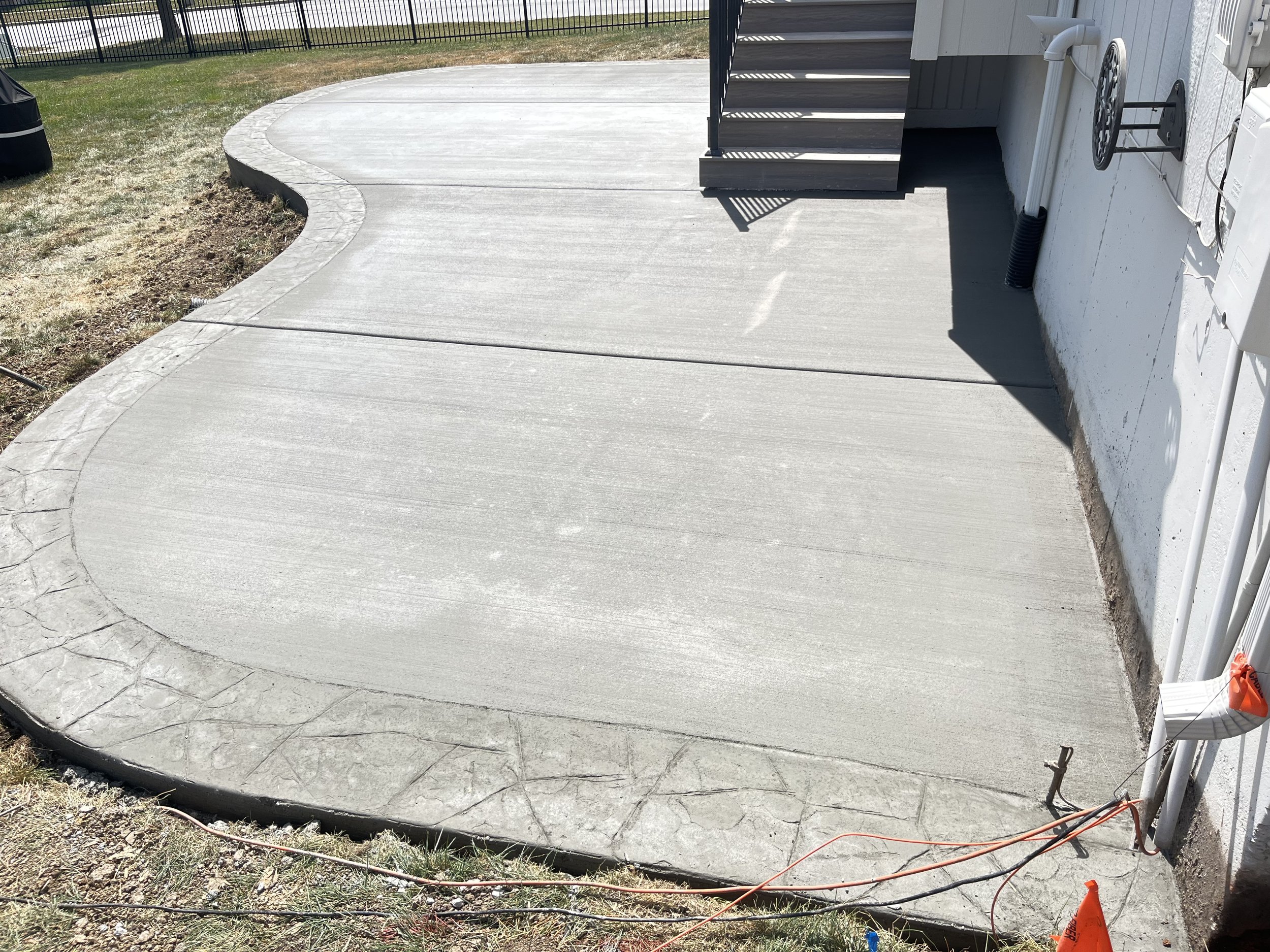 Newly poured concrete patio with a rounded edge, attached to stairs leading up to a house with a white exterior wall, and construction safety cones and wires in the foreground.