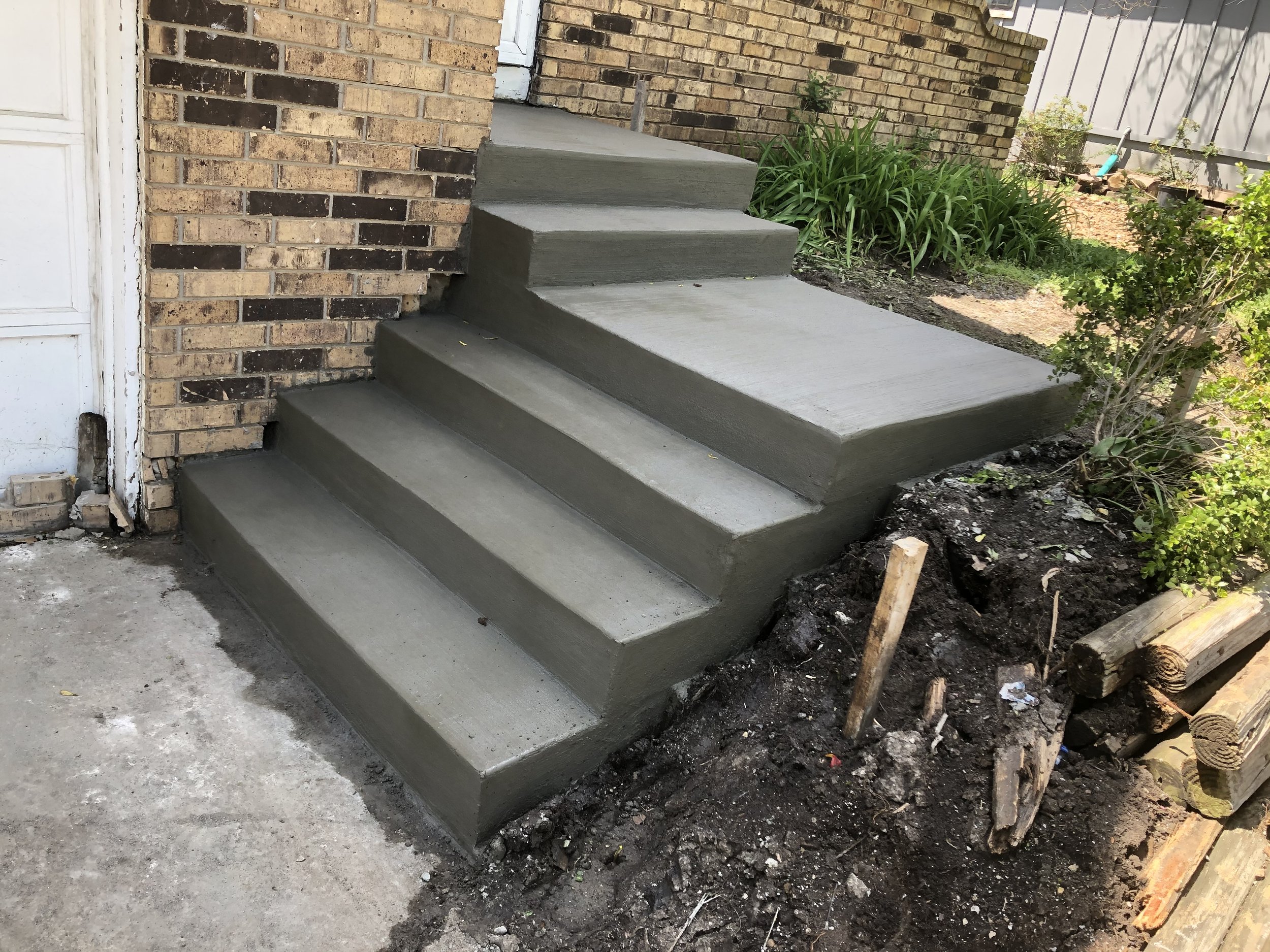 Newly finished concrete steps leading up to a house with brick exterior. The driveway and garden area are visible, with some construction debris and plant beds.