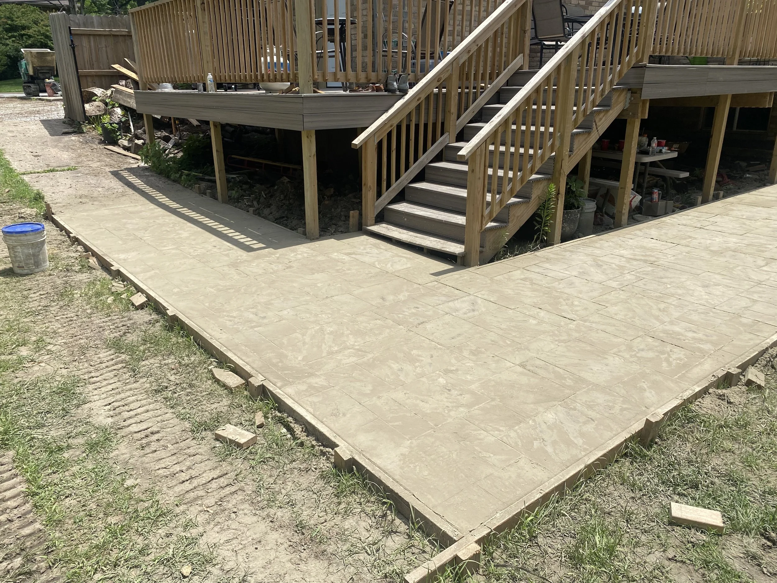 Newly constructed concrete patio with stamped pattern next to a wooden deck with stairs, under construction with some tools and materials nearby.