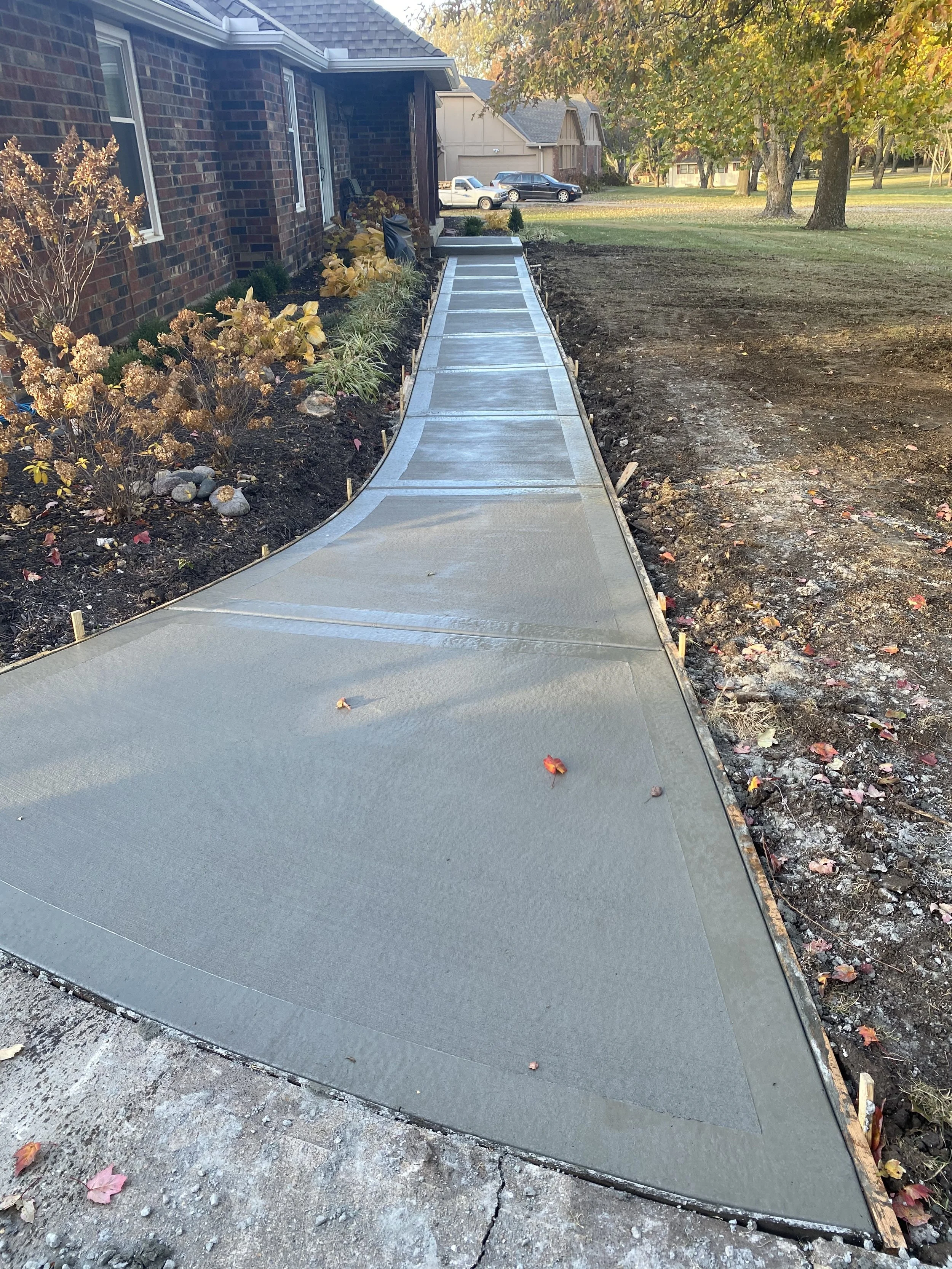Freshly poured concrete sidewalk in front of a brick house with landscaping and trees.