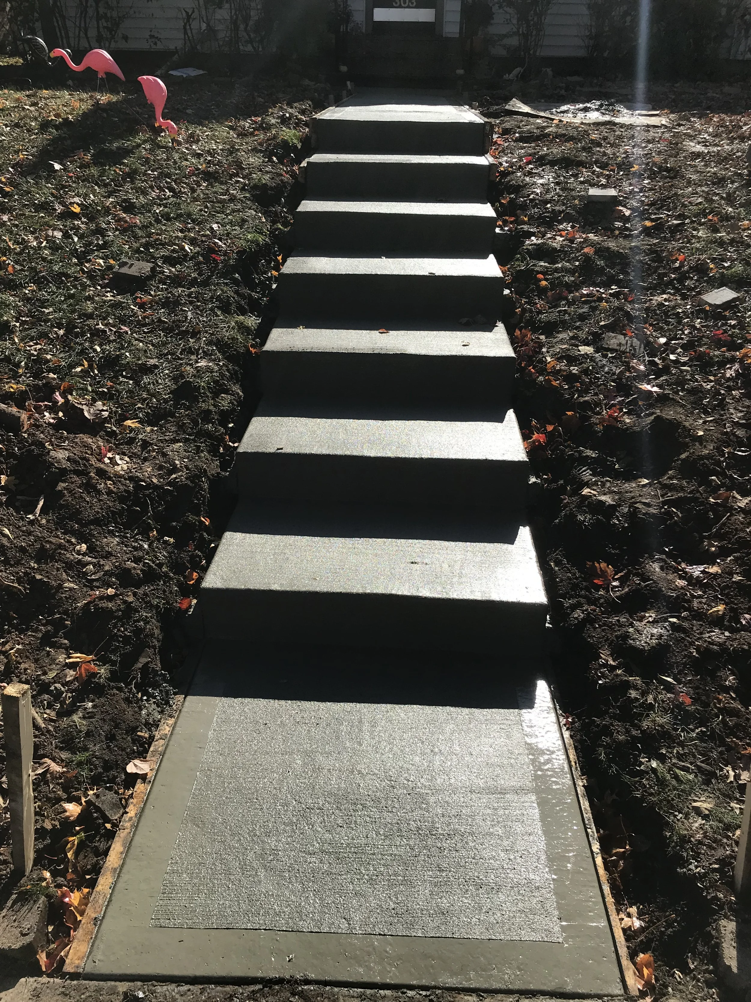 Newly poured concrete walkway with steps leading to a house entrance, flanked by soil with some scattered leaves and a pink flamingo lawn decoration.