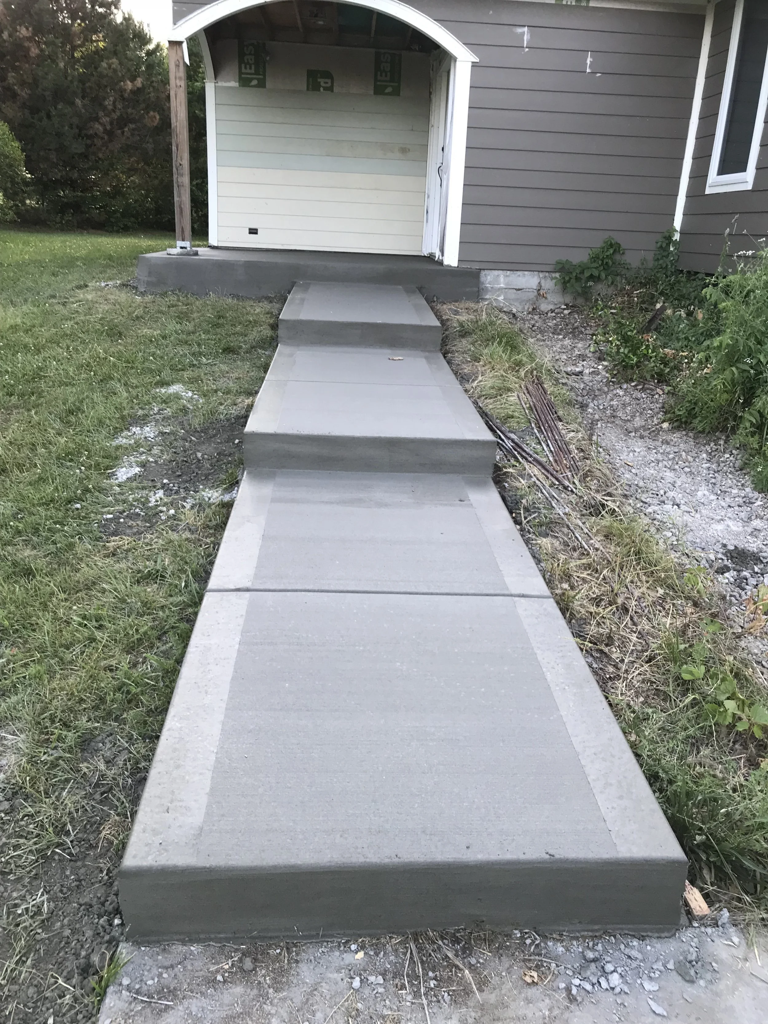 Newly poured concrete walkway leading to a house with a small porch under construction. The walkway has steps and is surrounded by grass and dirt.