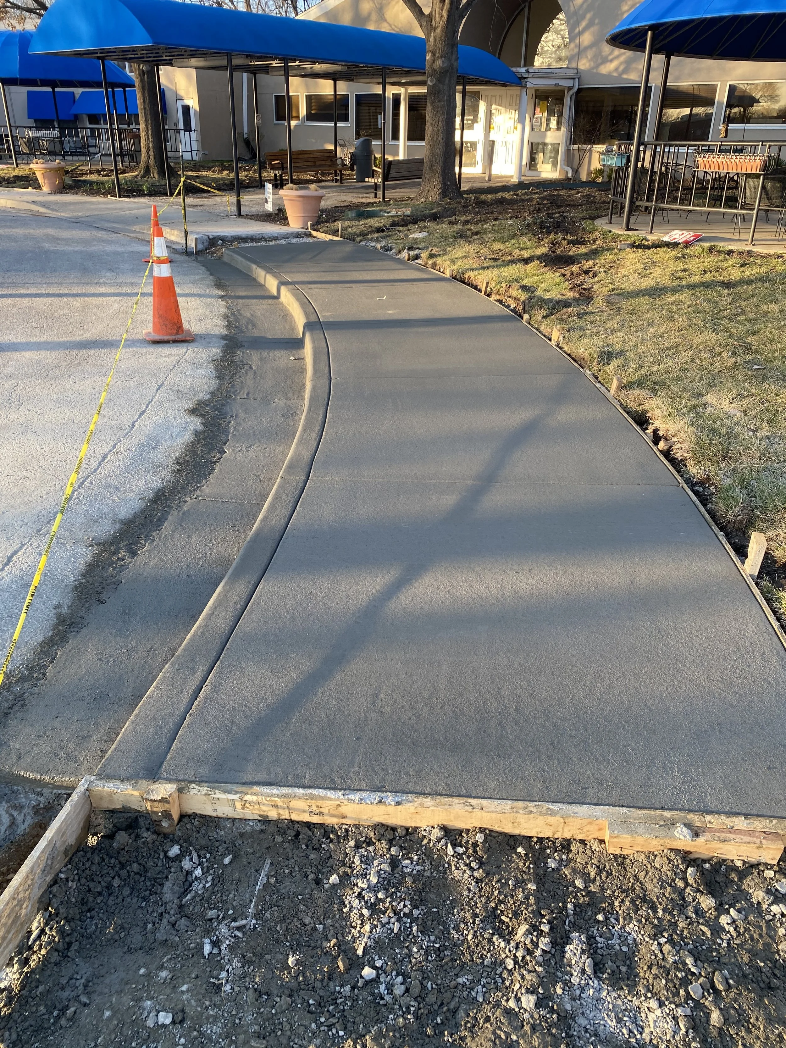 Freshly poured concrete sidewalk with a slight curve, bordered by wooden forms, near a building with blue awnings and outdoor seating area, with a traffic cone and caution tape nearby.