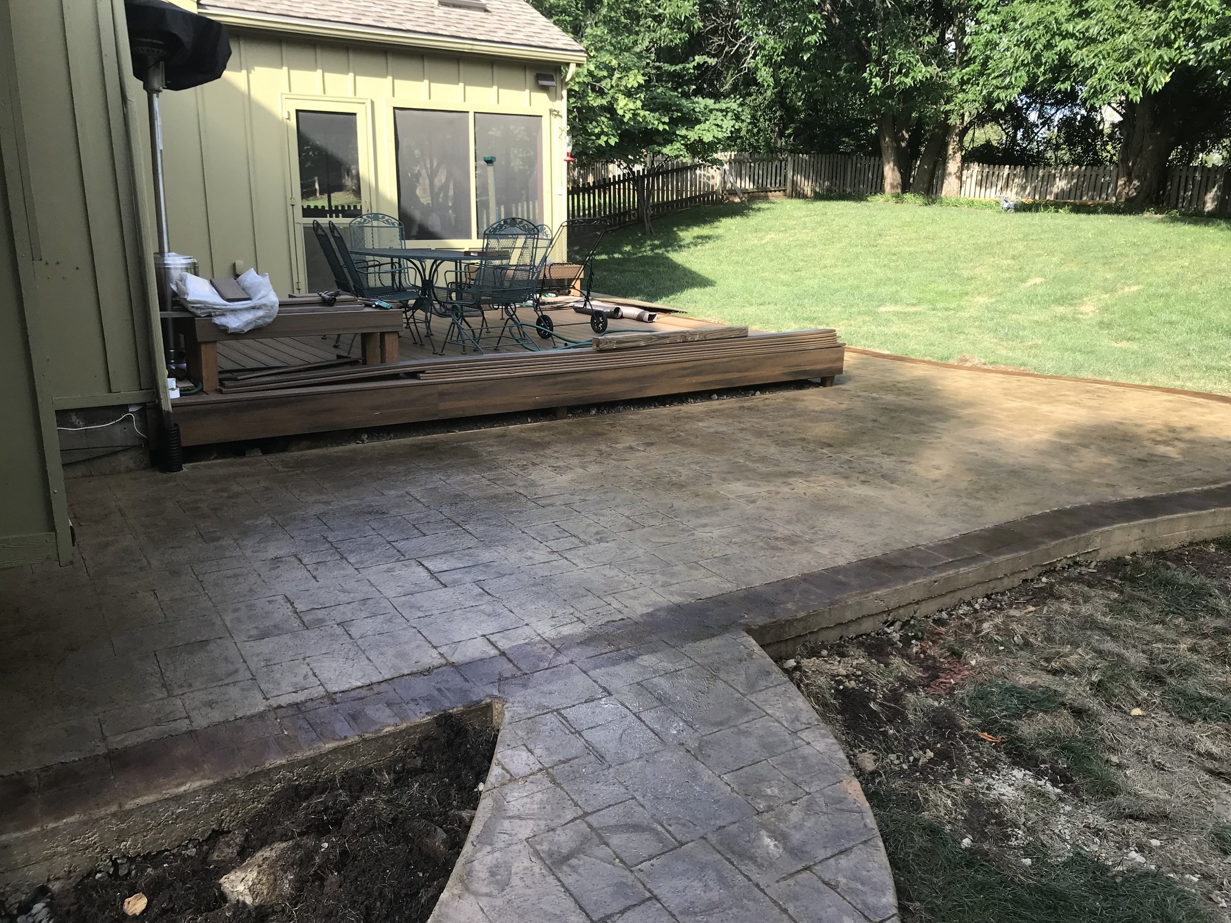 A backyard deck area with outdoor furniture, including a table and chairs, adjacent to a grassy lawn, with a small tree and a wooden fence in the background, under bright sunlight.