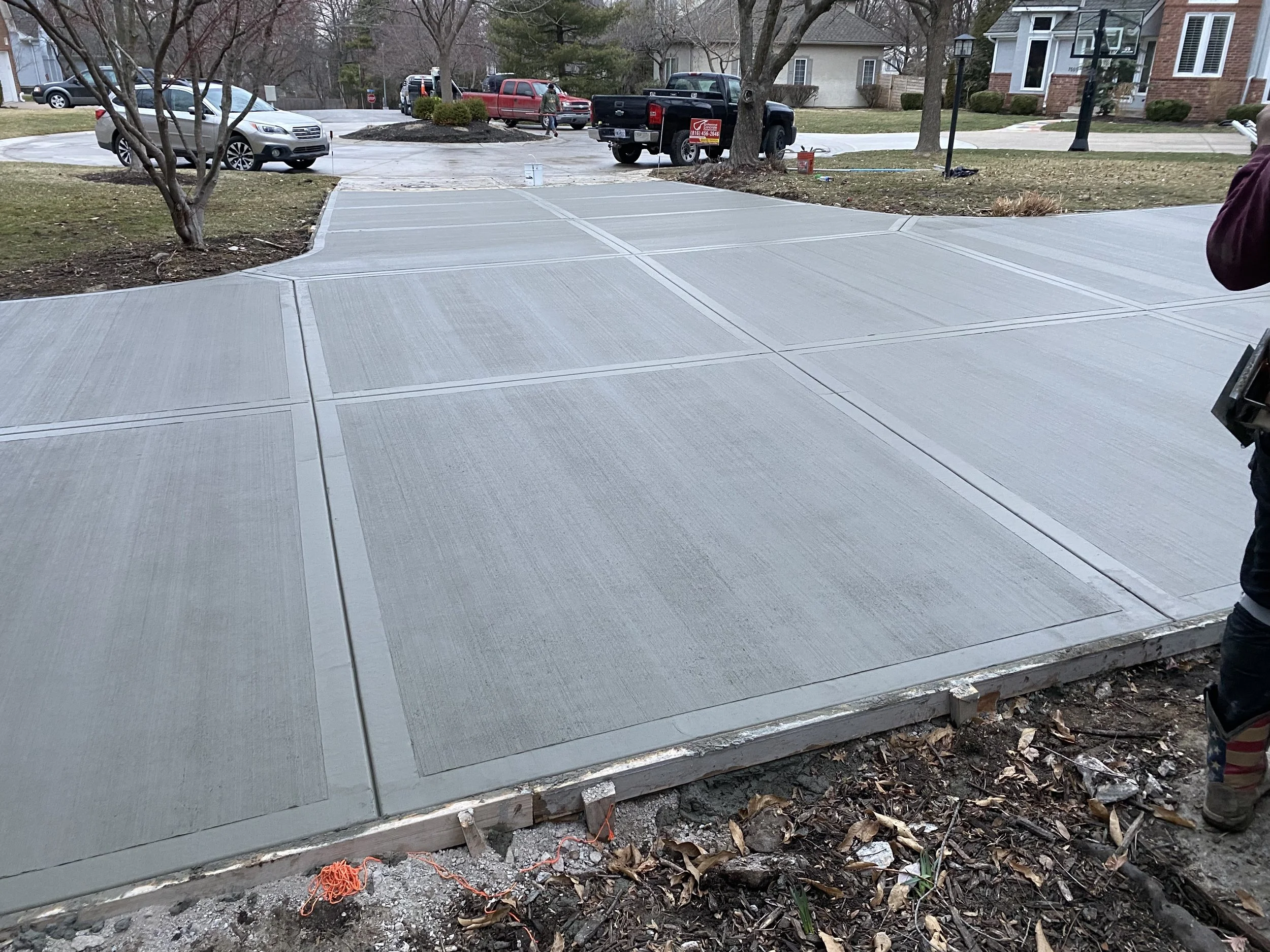 Freshly poured concrete sidewalk with expansion joints, surrounded by dirt and fallen leaves, in a residential area with houses, trees, and parked cars in the background.