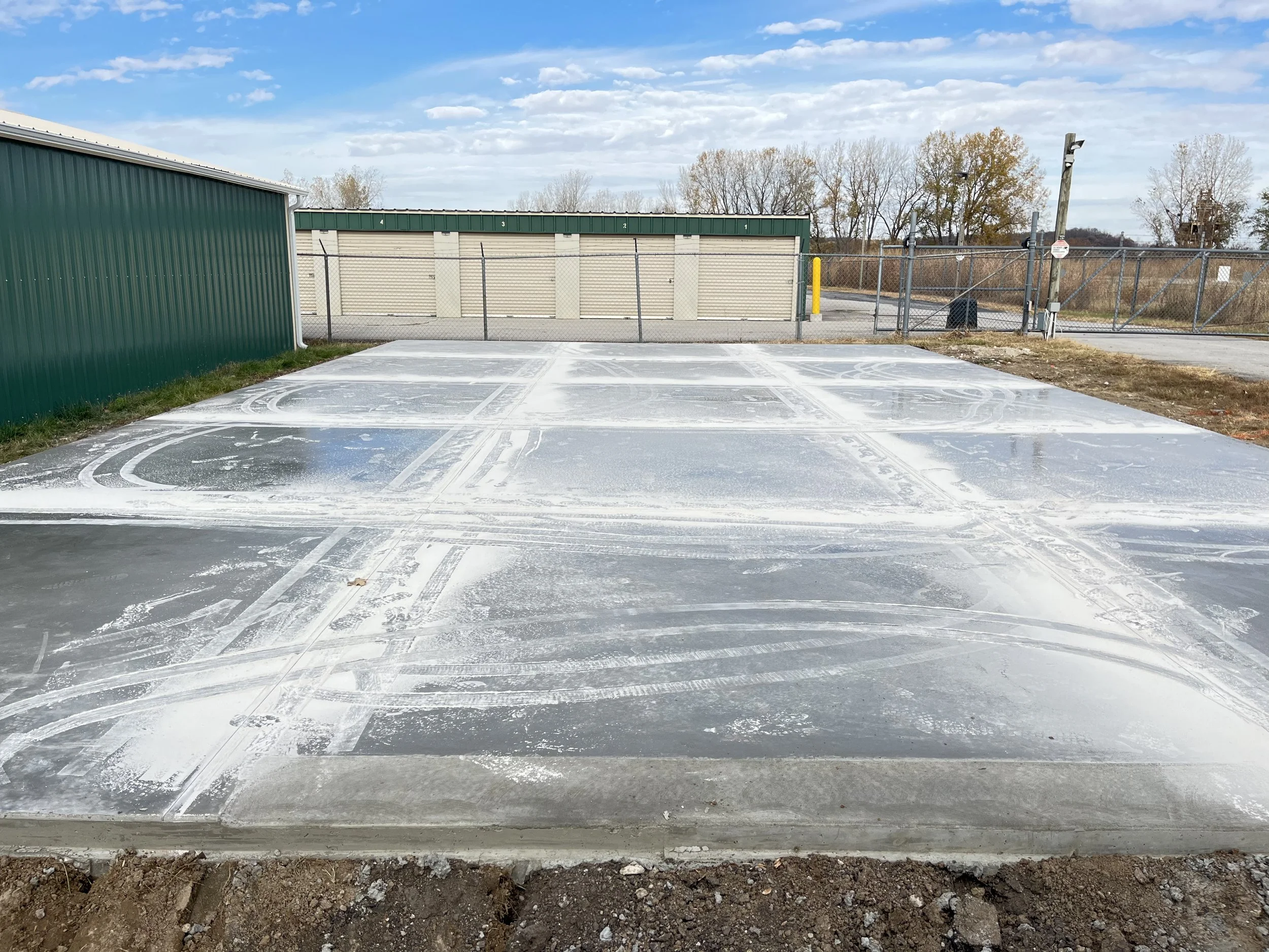 Empty parking lot with frost and tire tracks, surrounded by green and beige storage buildings, with a chain-link fence and trees in the background under a partly cloudy sky.
