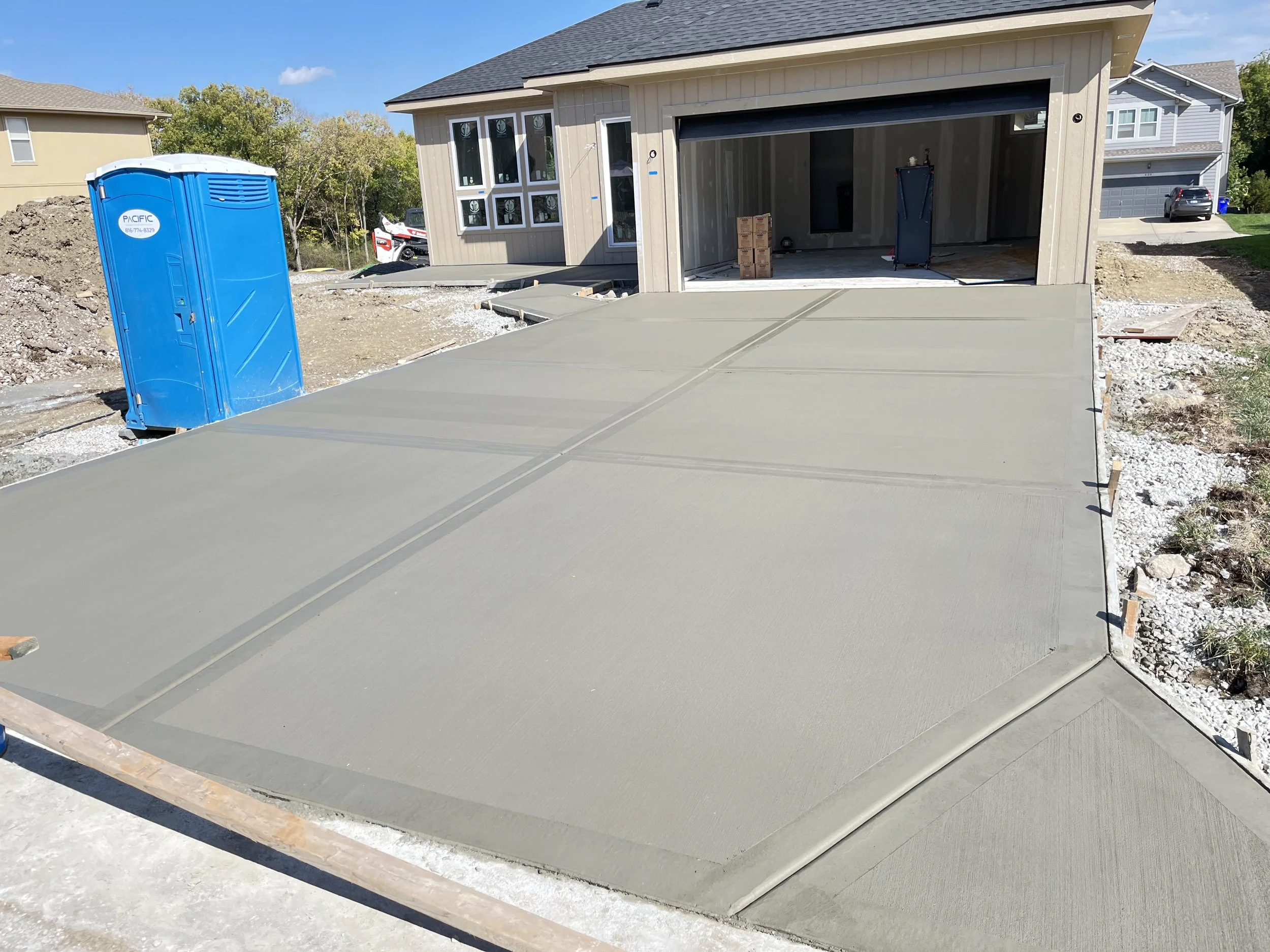 Freshly poured concrete driveway in front of a new house under construction, with a blue portable toilet on the left and construction materials inside the open garage.