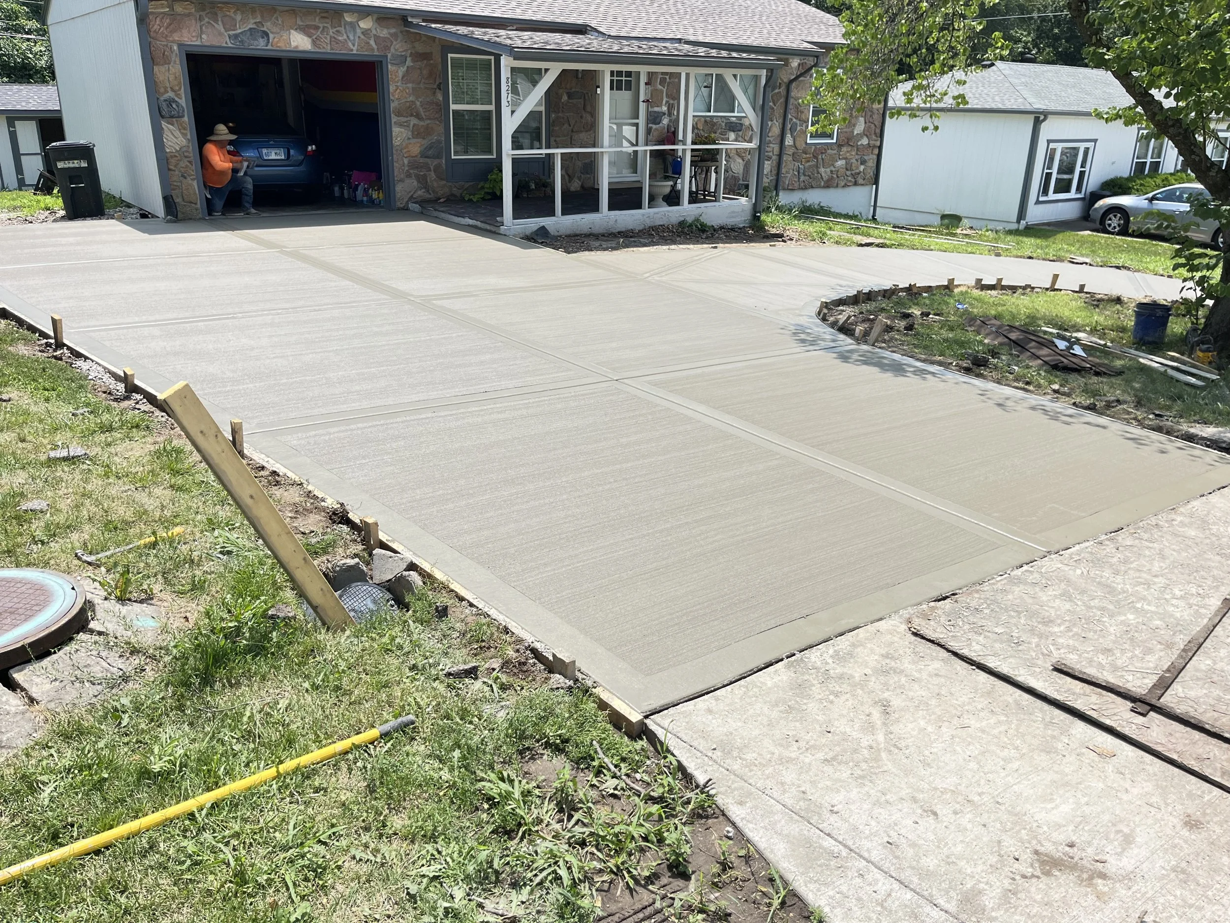 Newly poured concrete driveway in front of a house, with construction materials nearby and a person sitting in a garage.