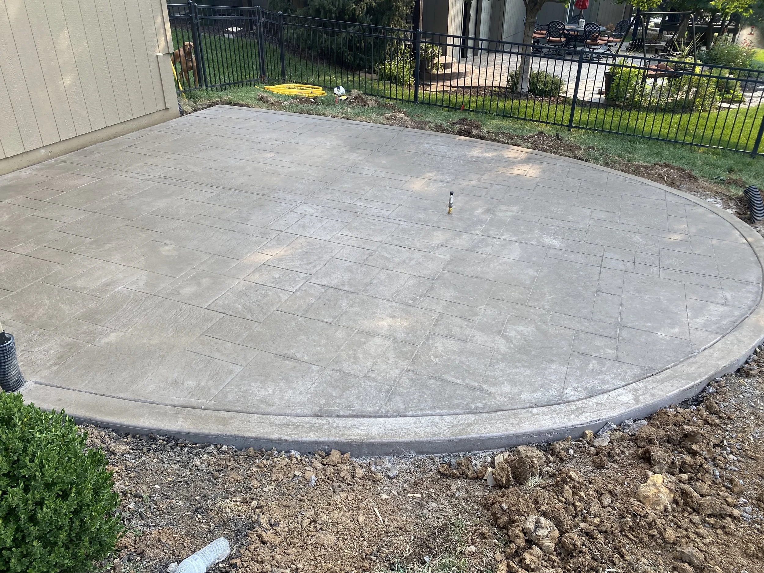 Newly paved concrete patio with decorative stamped design, surrounded by dirt and a small shrub in the foreground, with a black metal fence and landscaped yard in the background.