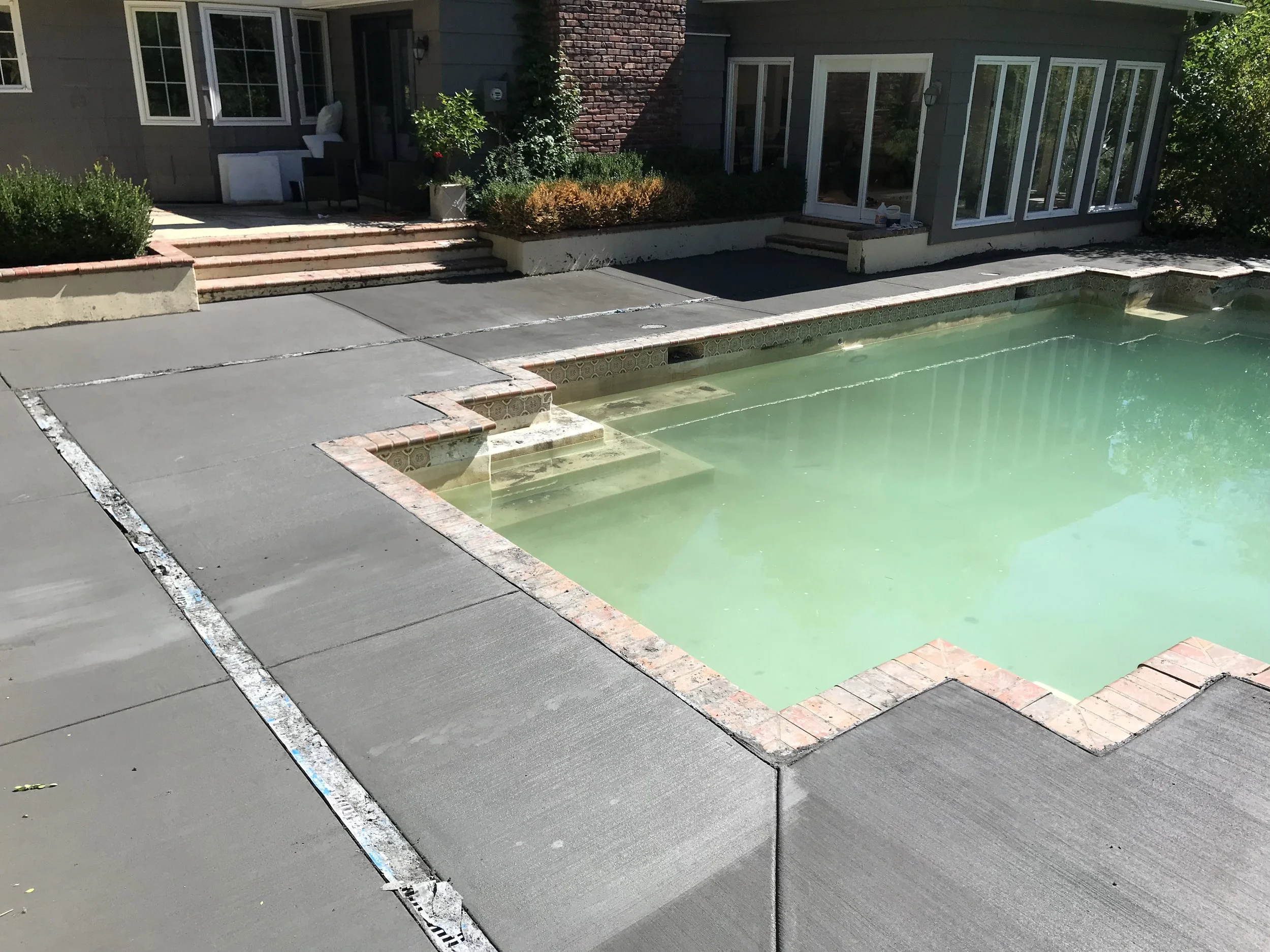 A swimming pool with light green water surrounded by a concrete deck and brick edging, with steps leading into the pool, located behind a backyard house with large windows and sliding glass doors.
