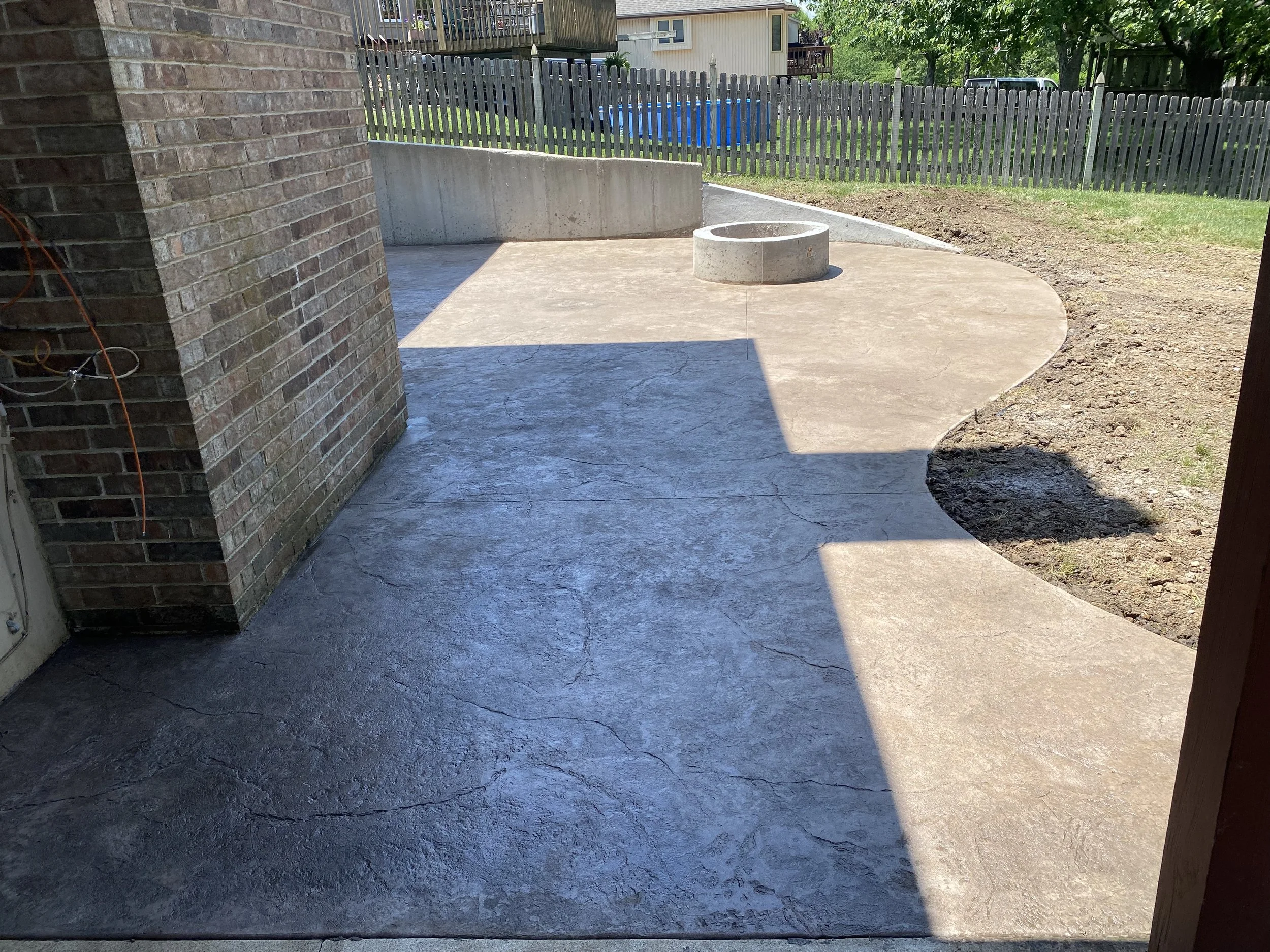 Concrete patio with a curved edge, brick wall on the left, a circular concrete fire pit, a wooden fence, green grass, and trees in the background.