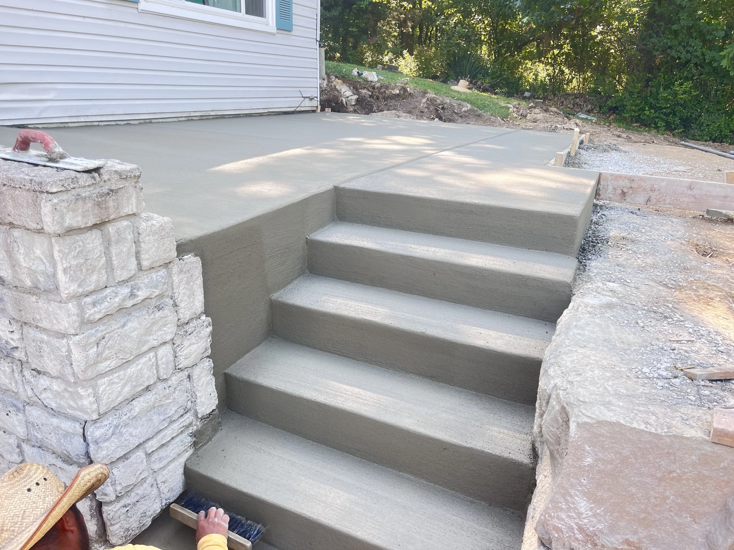 New concrete steps and patio area with a stone retaining wall, outside a house with white siding and blue shutters, surrounded by greenery.