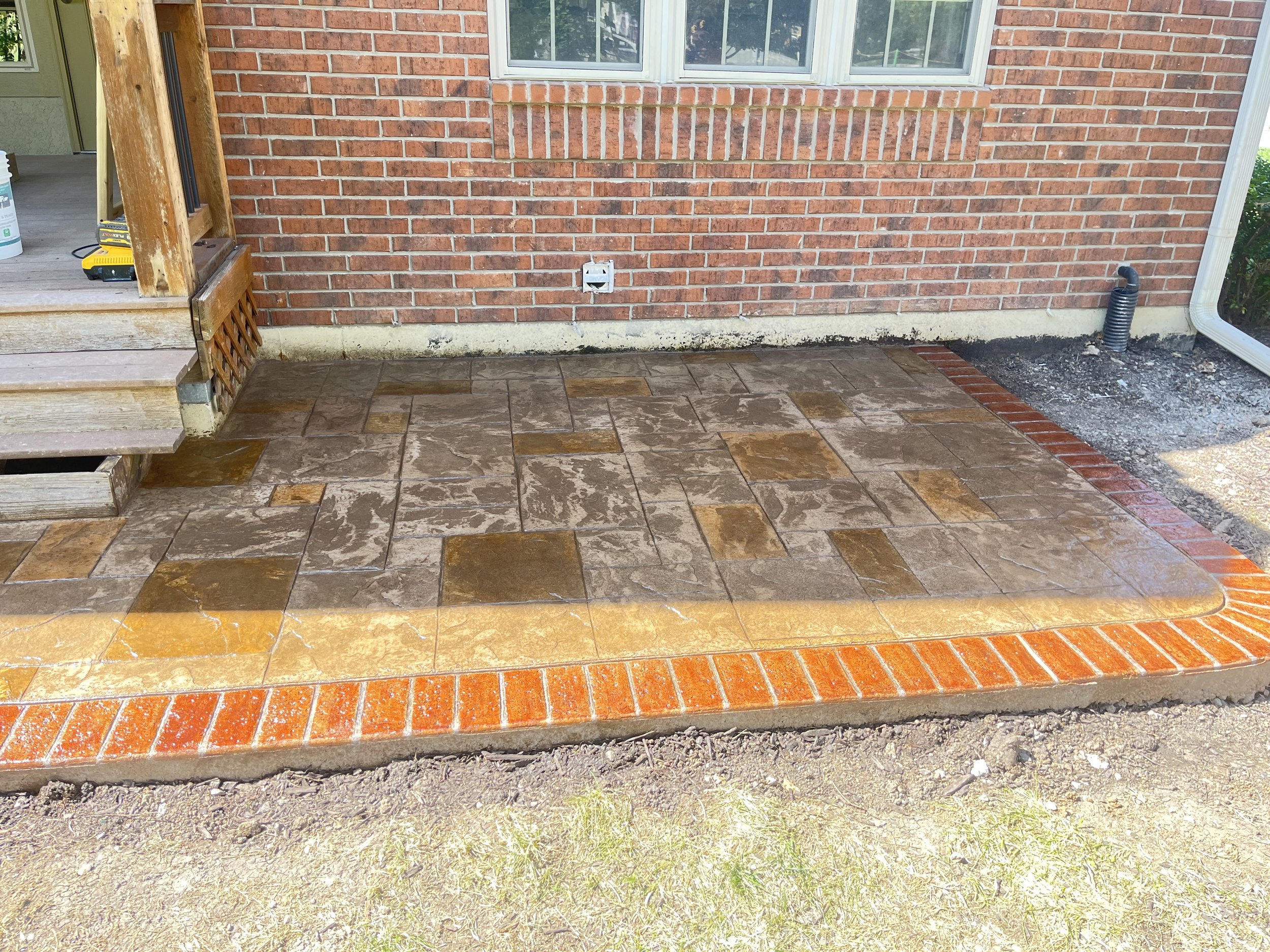 Newly installed outdoor brick patio with stamped concrete, surrounded by dirt and a brick house wall with windows.