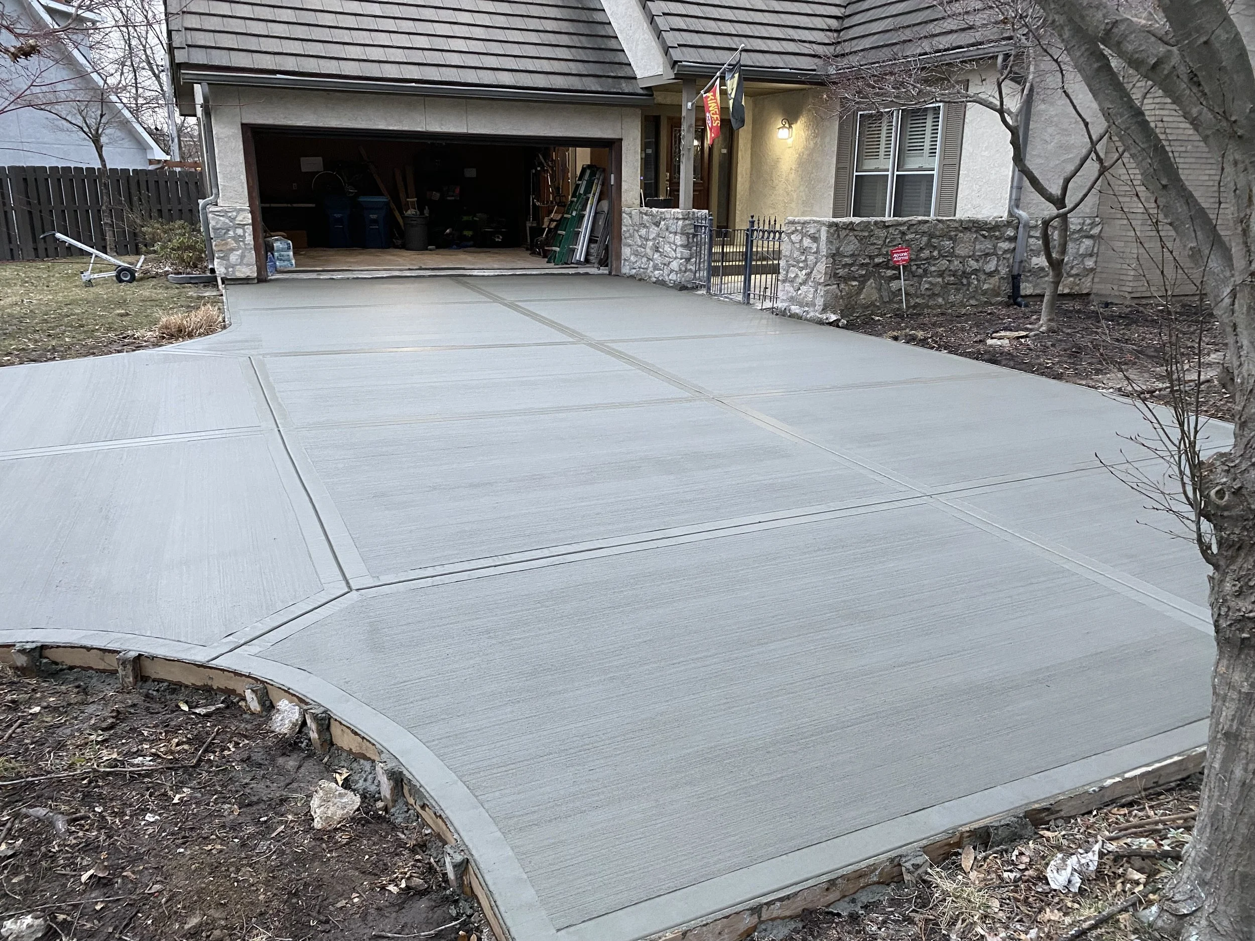 Newly poured concrete driveway in front of a house with an open garage, trees, and landscaping.