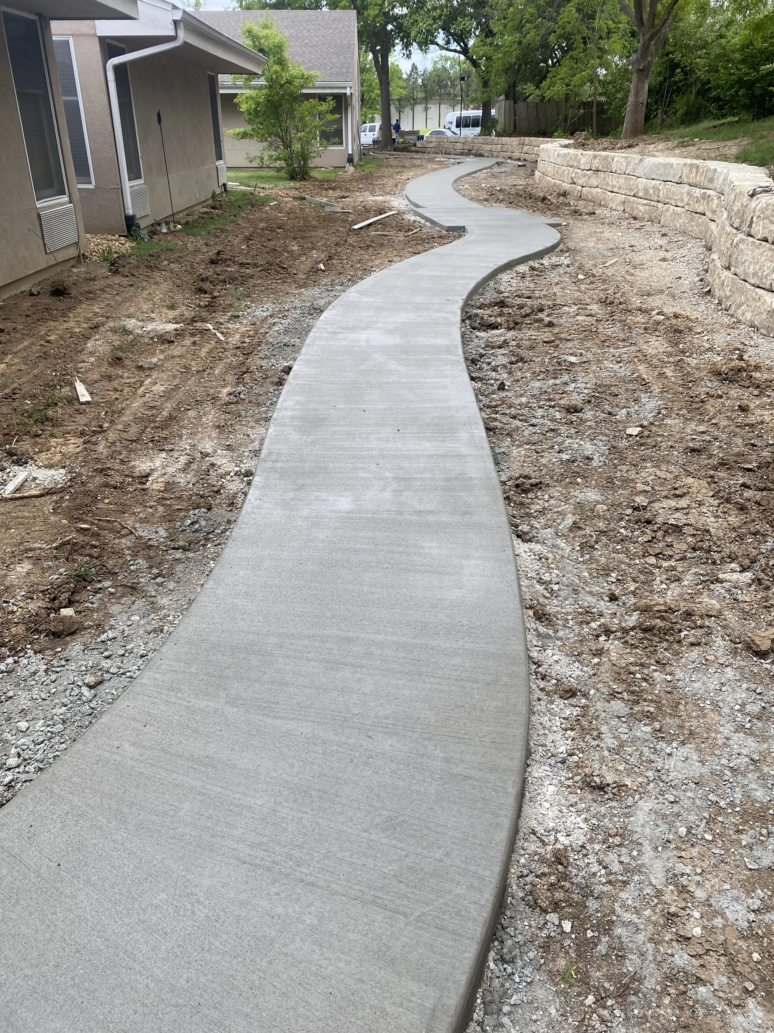 Newly constructed concrete sidewalk with a winding path runs adjacent to a house, supported by a retaining wall made of stacked stones on the right, with trees and parked vehicles visible in the background.