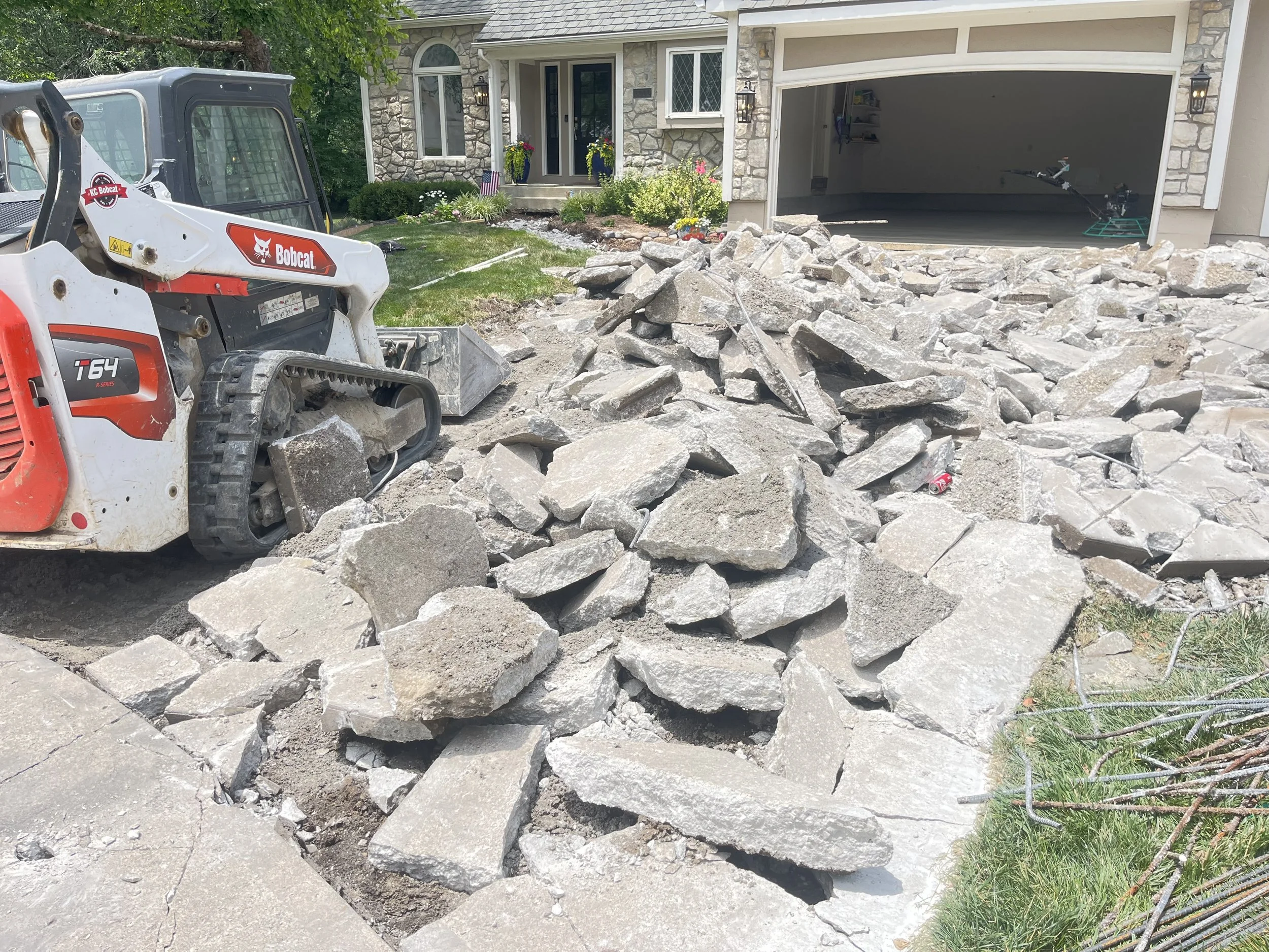 A driveway is being demolished with broken concrete and rocks, a skid-steer loader is nearby, in front of a house with a stone facade and a garage door open.