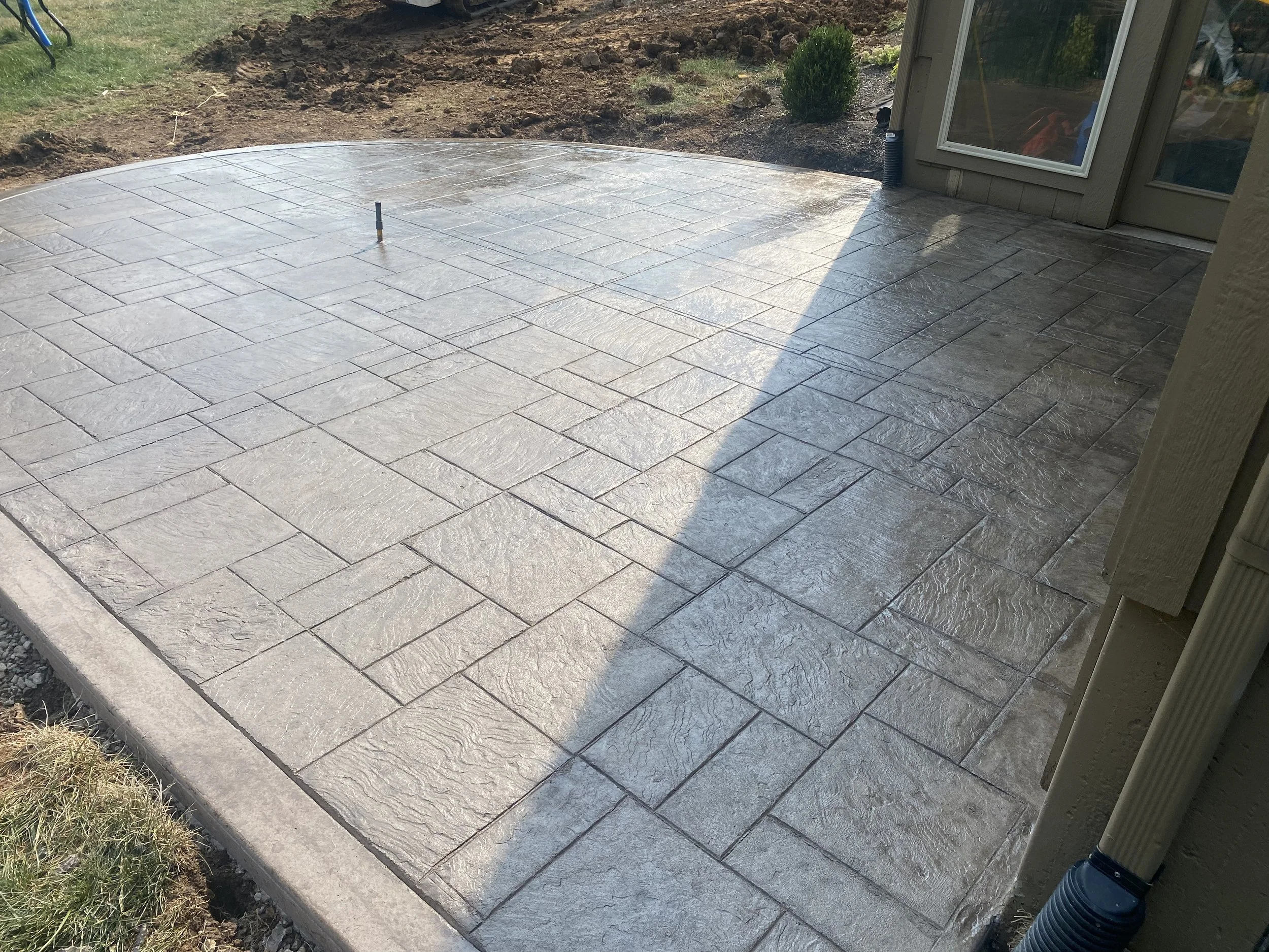 Freshly laid concrete patio with stamped pattern, surrounded by garden soil and a small shrub, next to a house with a sliding glass door.