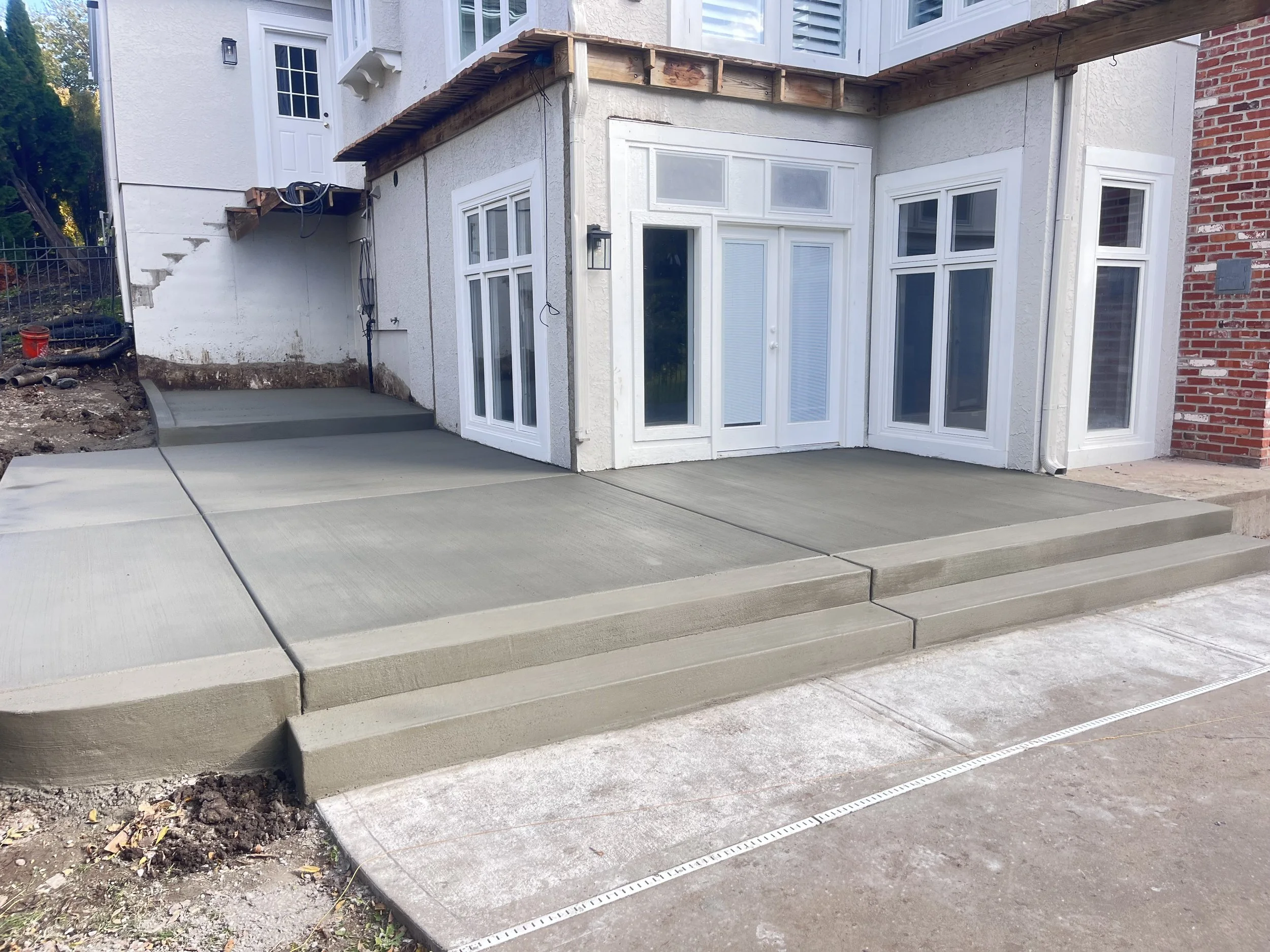 Newly poured concrete patio and steps outside a house with large white framed glass doors and windows, construction in progress with some dirt and bricks visible on the side.