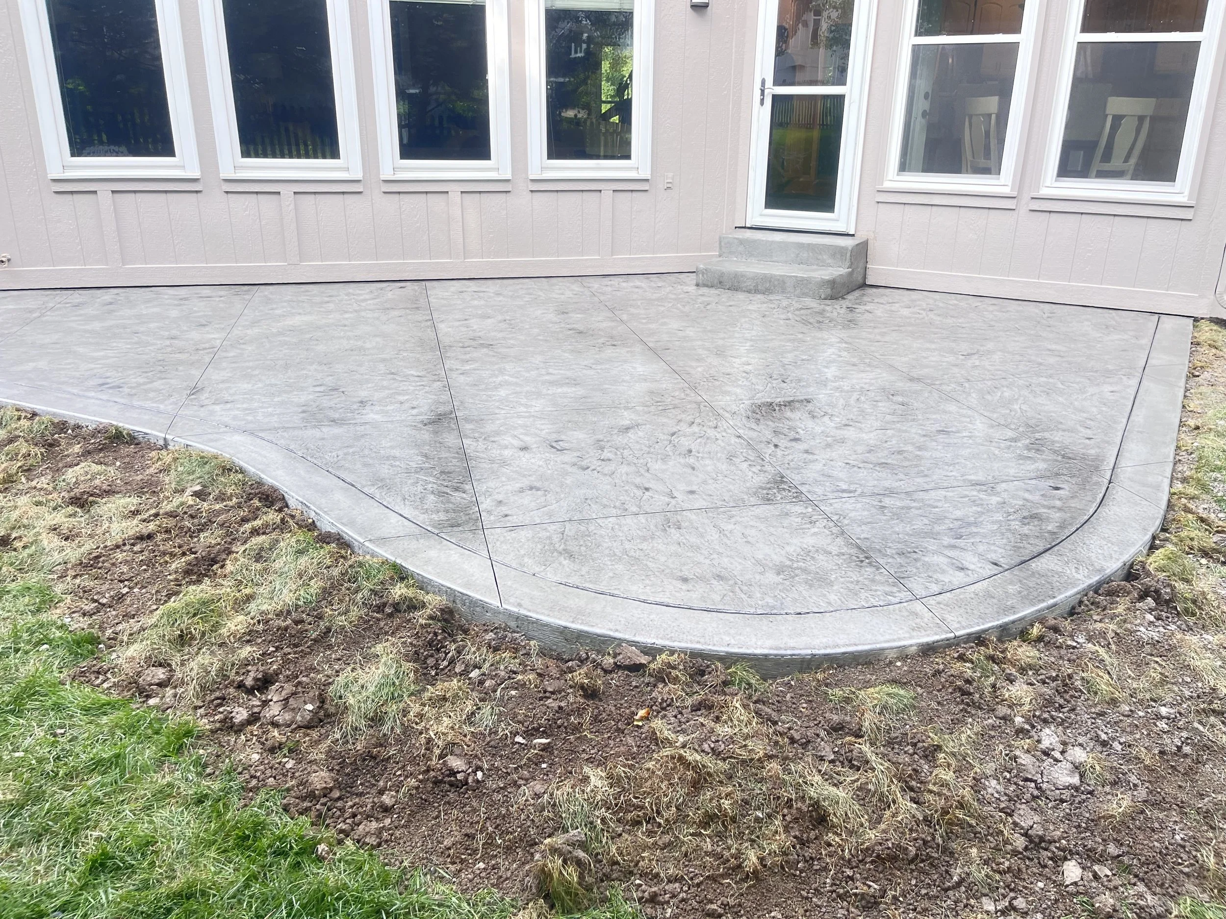 Concrete patio with stamped pattern, attached to the house with a door, surrounded by grass and soil.