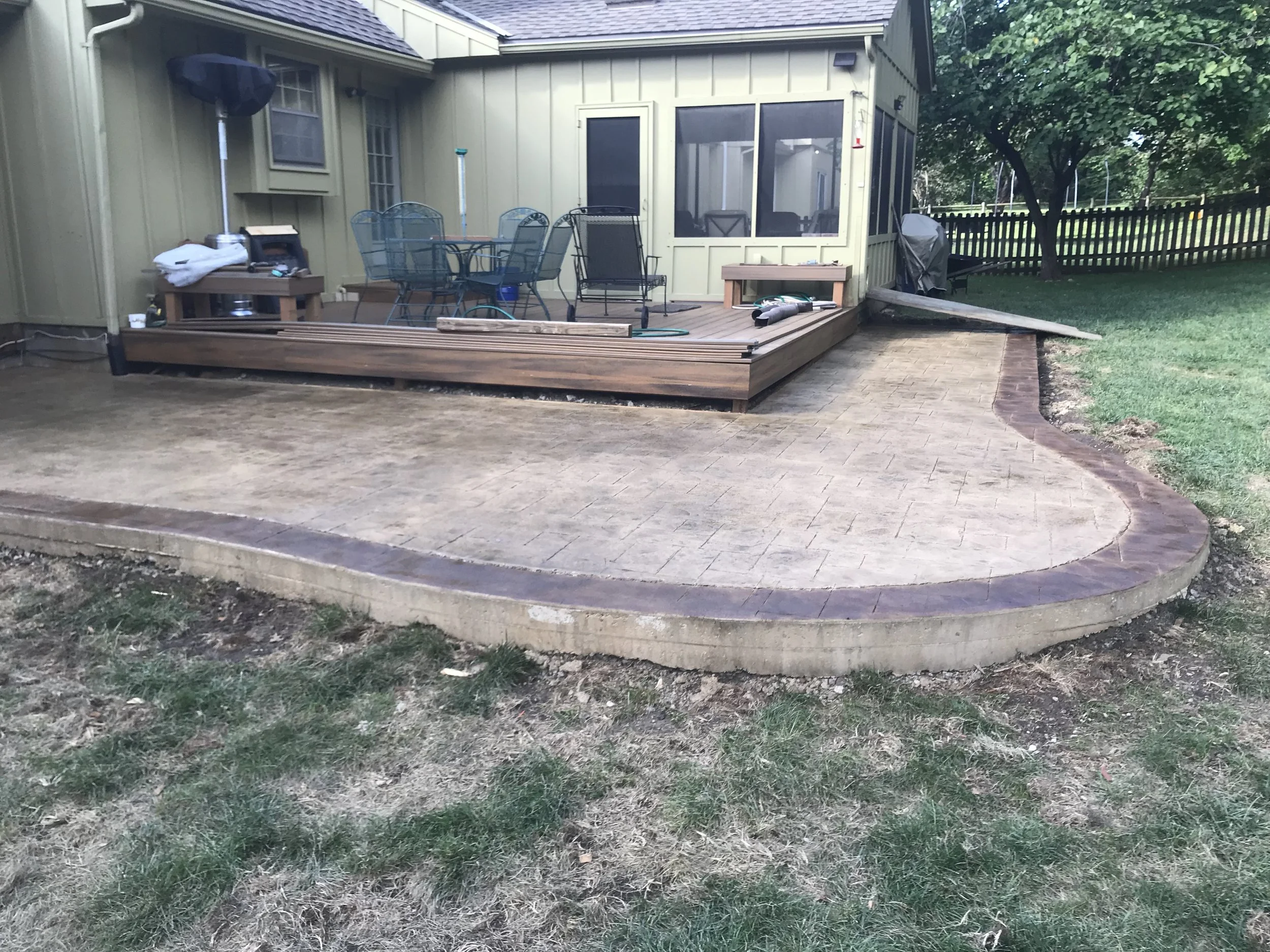Backyard with a newly built raised wooden deck, outdoor furniture, a barbecue grill, and a screened porch. The yard has a brick-paved patio area and grass, with a black fence and trees in the background.