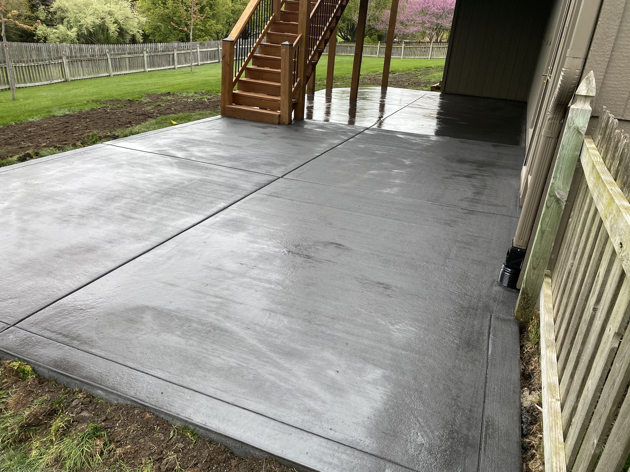 Freshly poured and smooth concrete patio with visible expansion joints, attached to a house with wooden staircase leading up from the patio, surrounded by a yard with grassy areas and a wooden fence in the background.