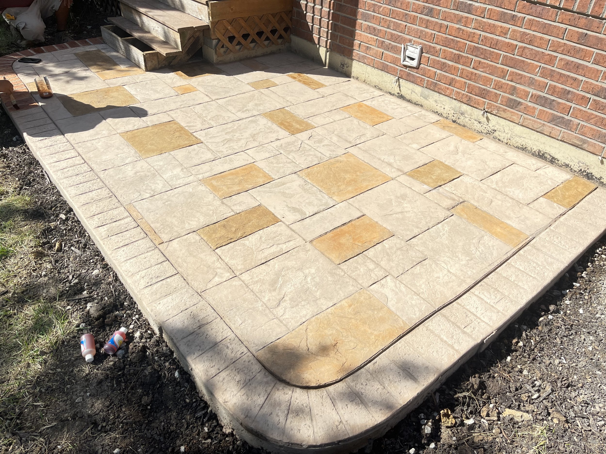 Newly constructed stone patio with beige and tan tiles adjacent to a brick house wall, with construction materials and tools nearby.