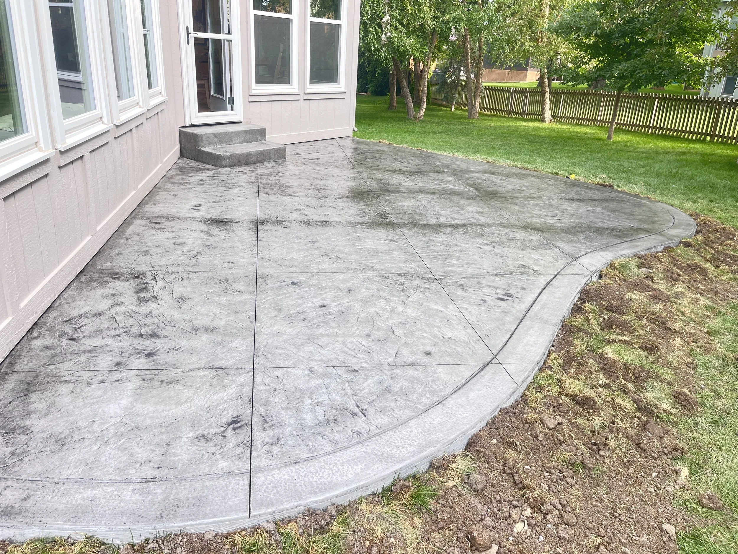 Concrete patio with a curved edge, adjacent to a house with multiple windows, and a grassy yard with trees in the background.