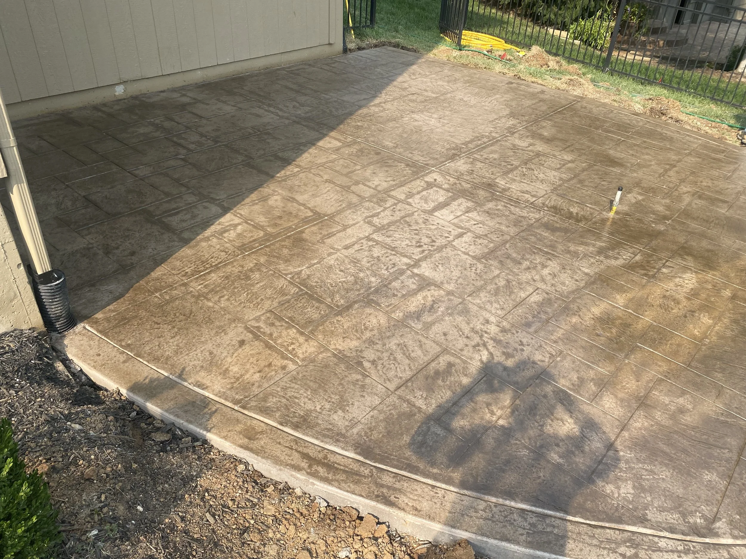 Freshly poured concrete patio with stamped pattern, partially wet, next to a house wall and a black metal fence with garden plants in the background.