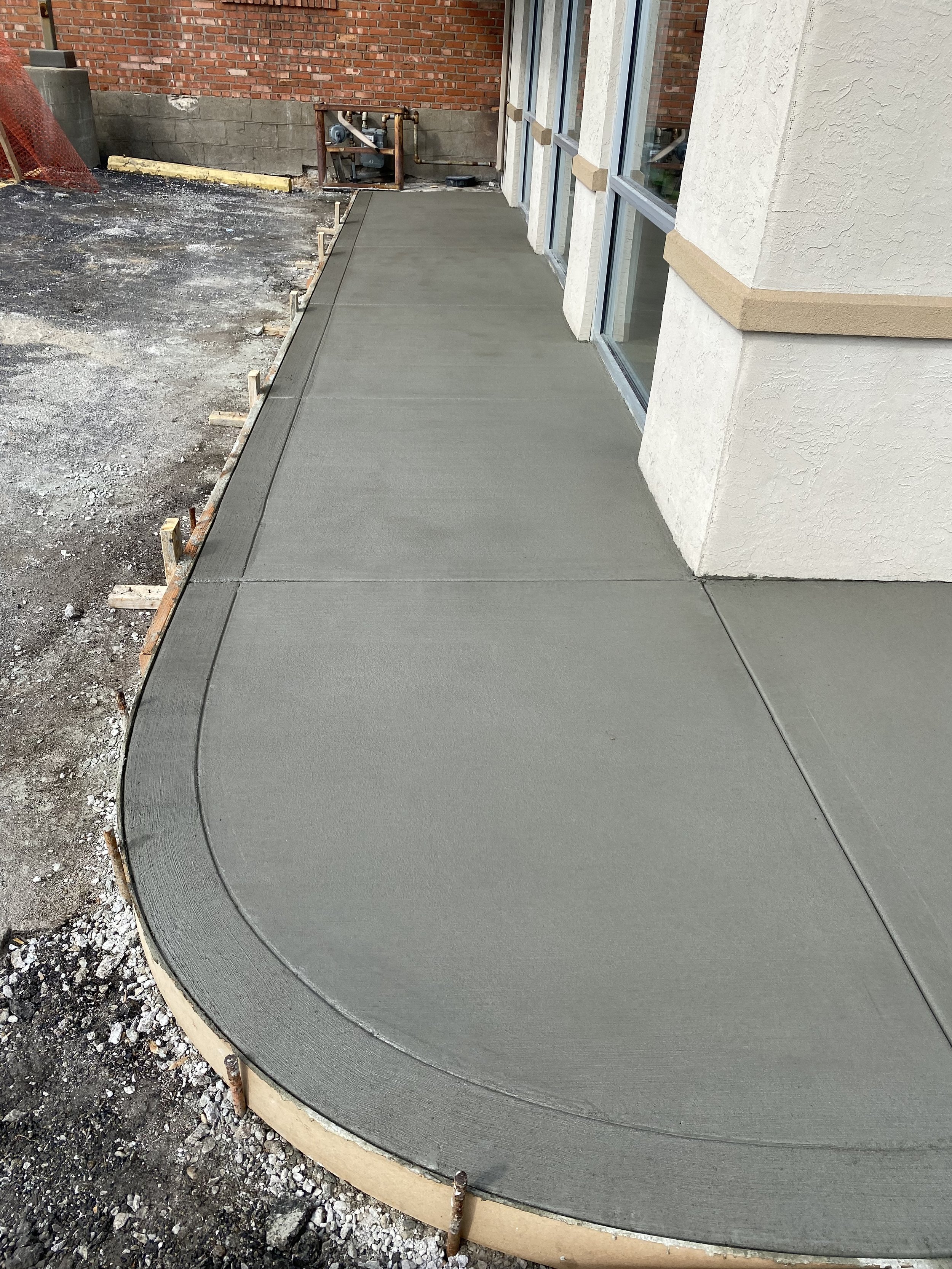 Freshly poured concrete on a curved sidewalk outside a building with windows and brick wall, with construction supports around the edge.