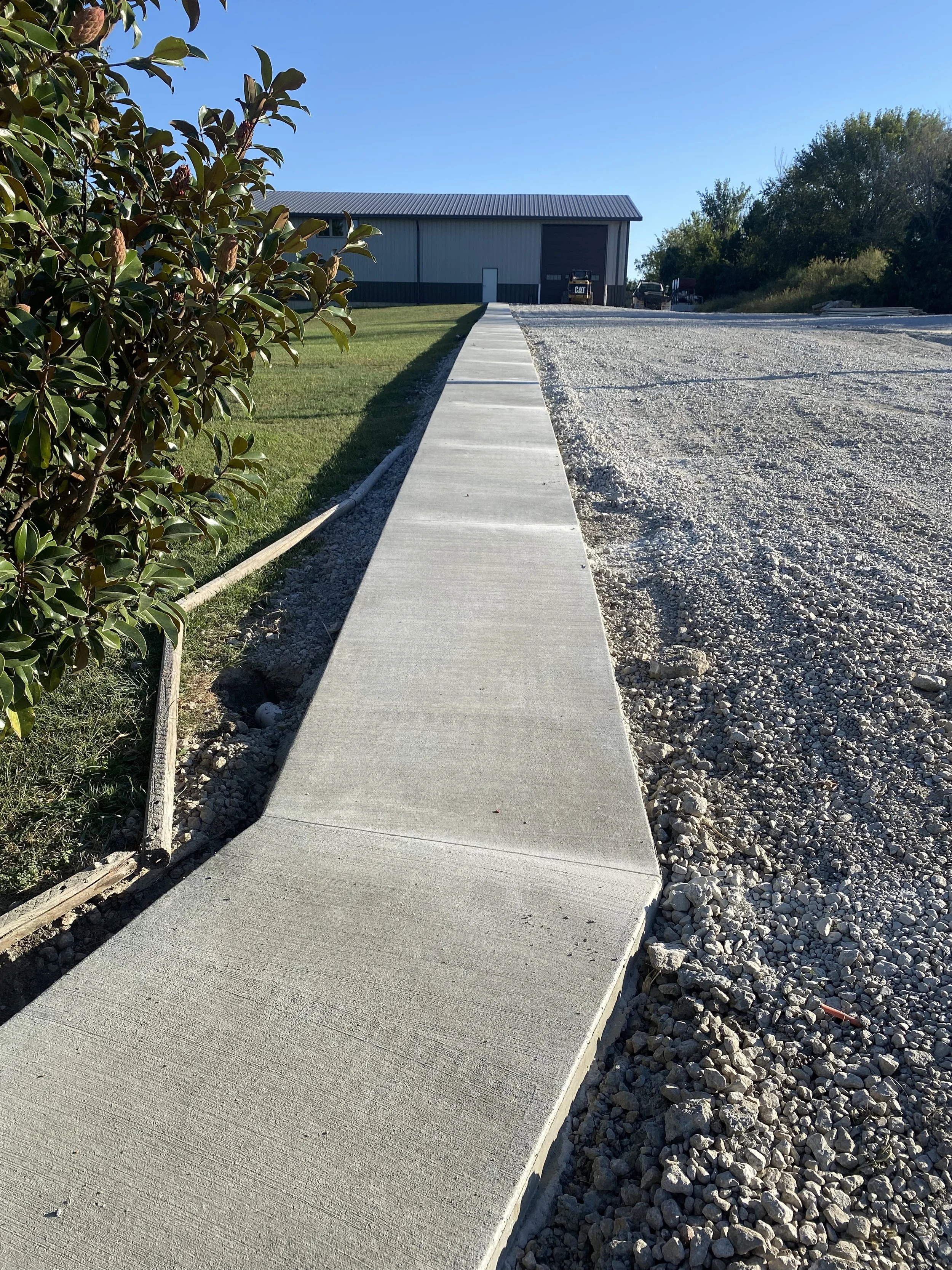 Concrete sidewalk running alongside a gravel parking lot, with a grassy area and bushes to the left and a large metal building in the background.