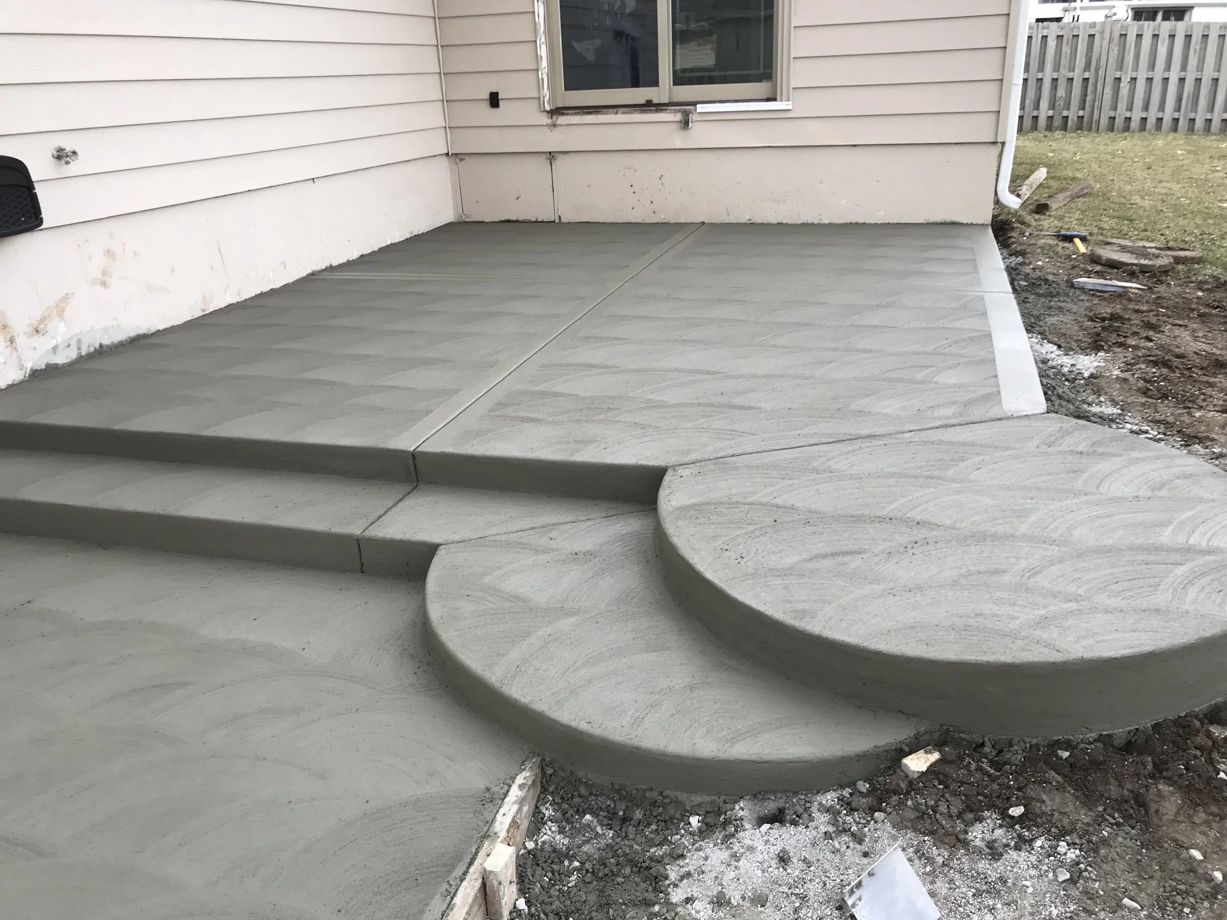 Newly poured concrete patio with curved steps outside a house, area surrounding the patio is under construction.