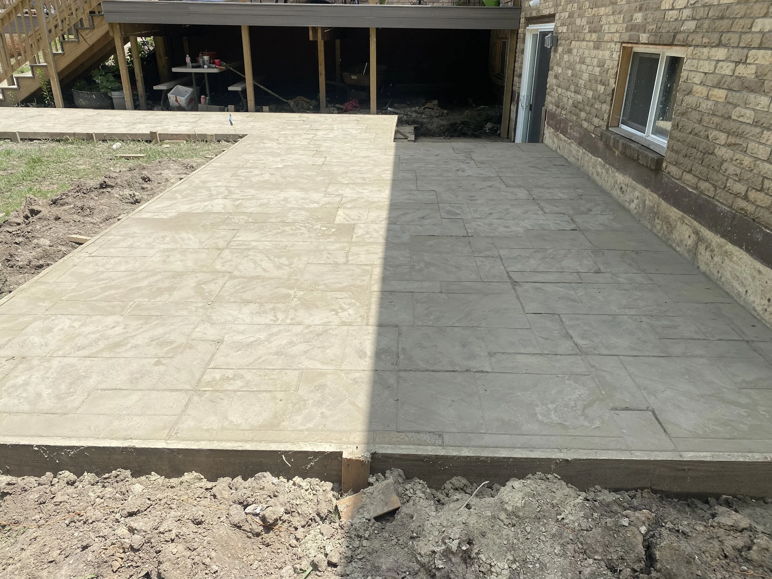 Newly paved concrete patio area with stone-like pattern next to a brick house, with some soil and dirt at the edges mingling with the paving.