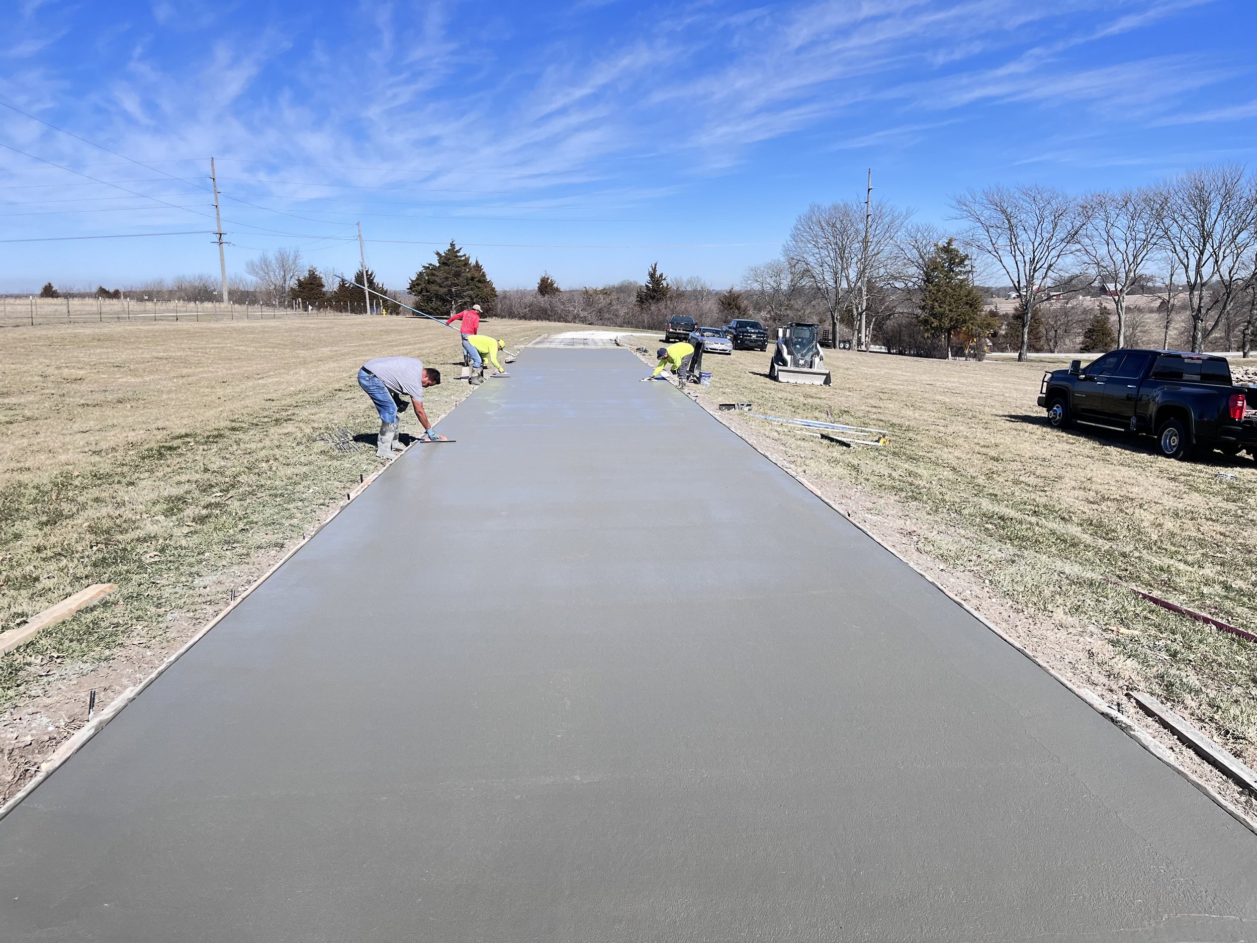 Construction workers pouring and smoothing concrete on a road in an open field on a sunny day.