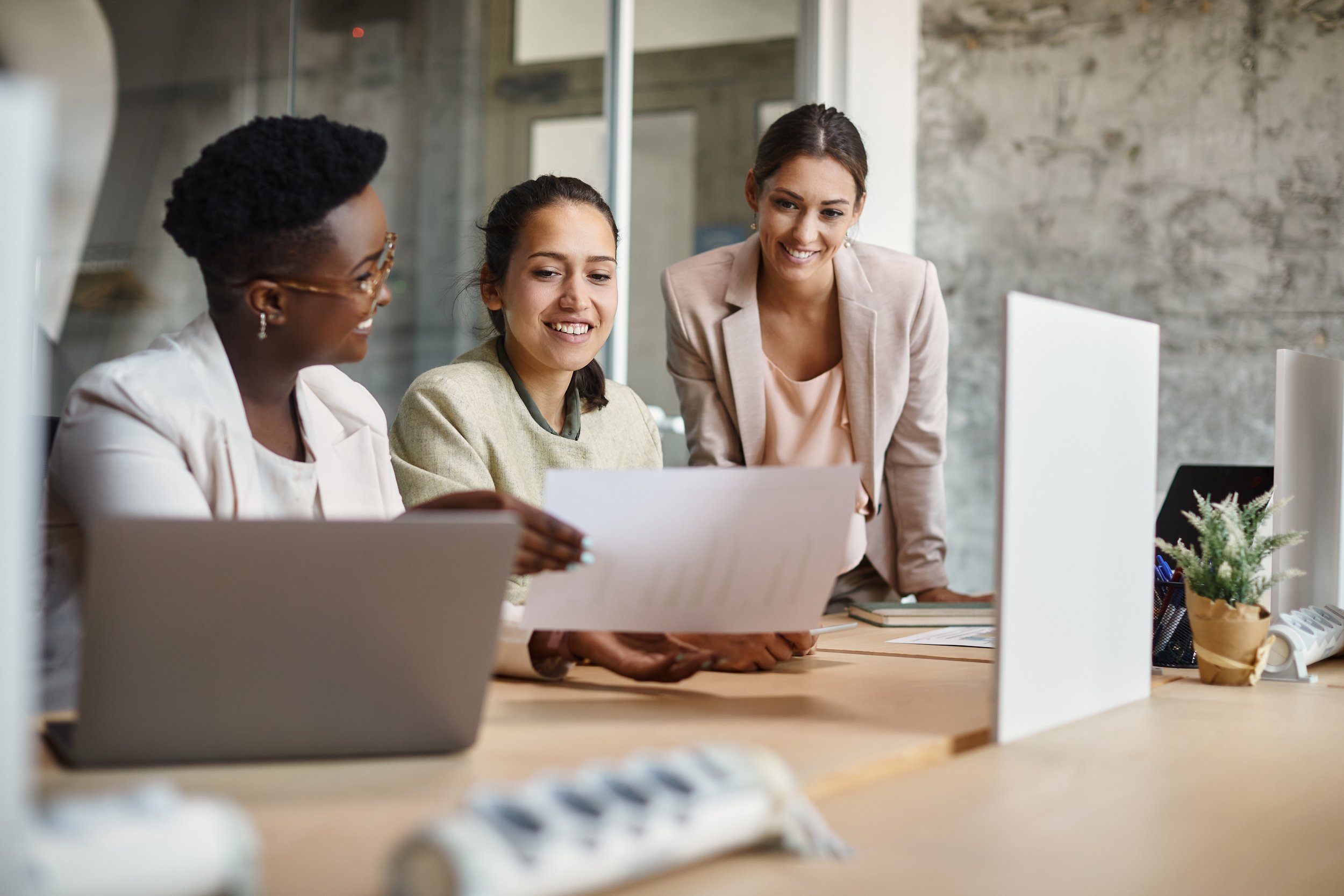 Three women in an office are looking at a document and smiling, one woman stands leaning over the desk.