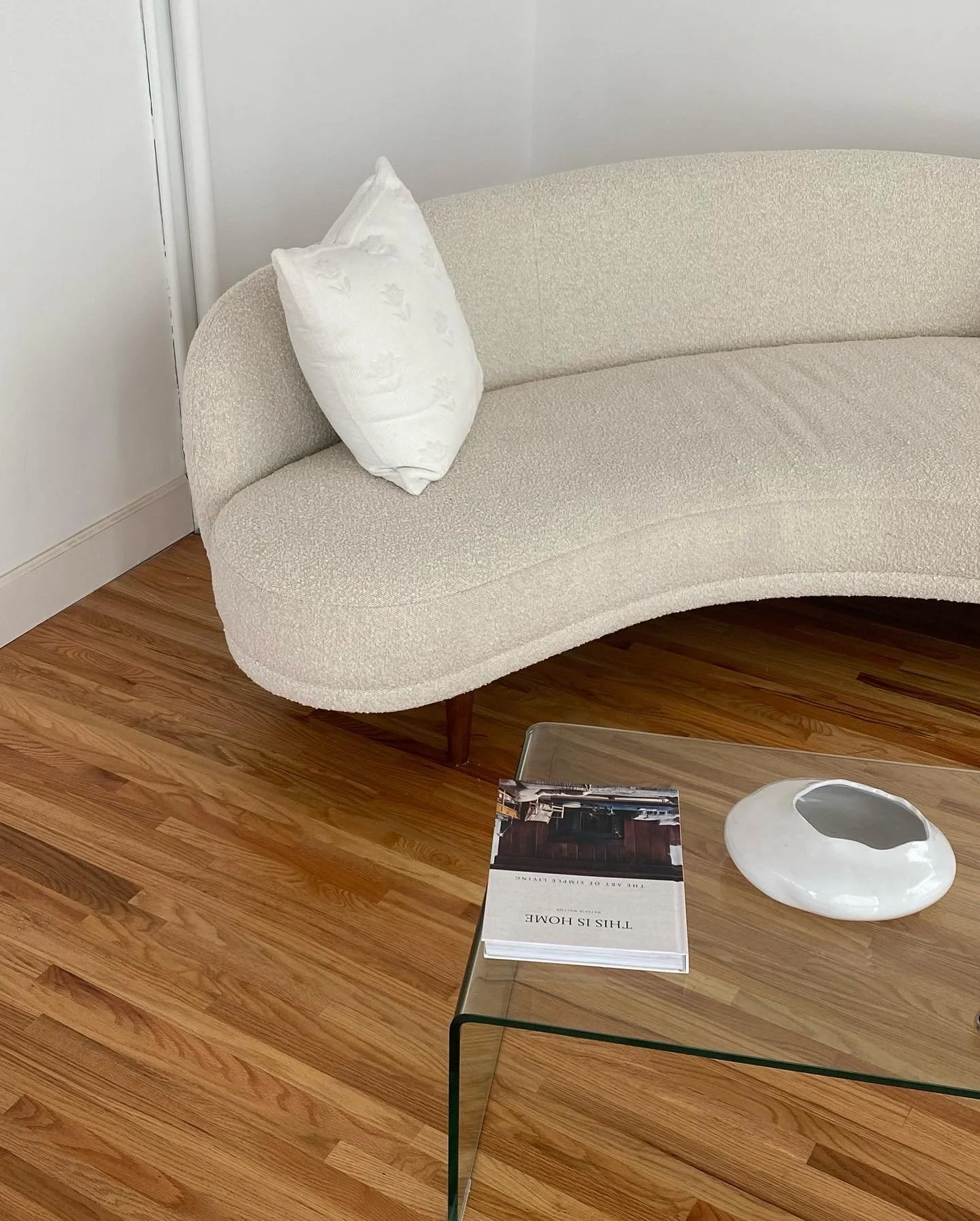 A beige curved sofa with a white pillow, a glass coffee table with a magazine titled 'This is Home' and a white ceramic decorative object, with hardwood flooring.