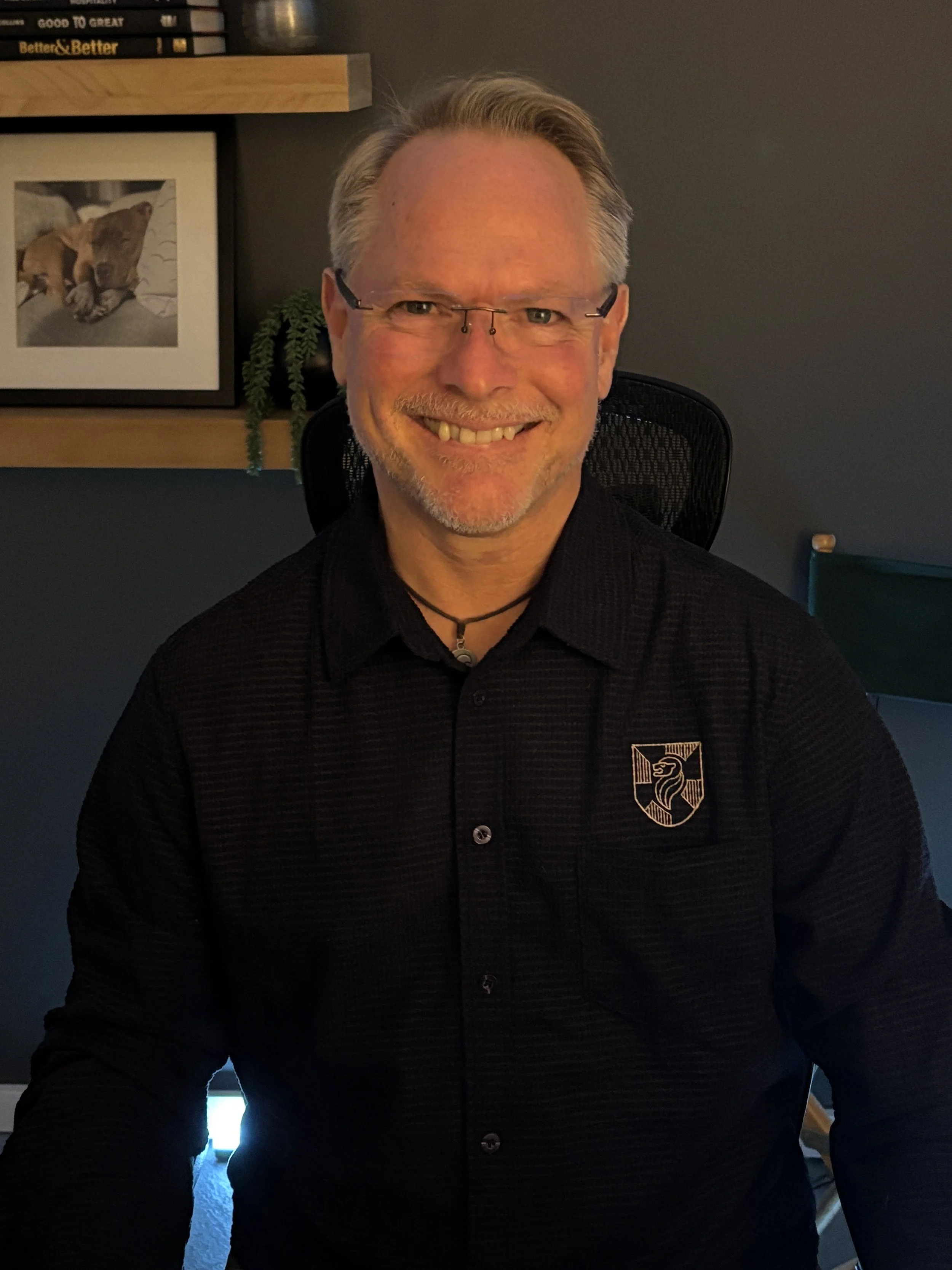A smiling man with glasses, short gray hair, and a beard, wearing a black collared shirt with a crest on the chest, sitting in a room with dark walls, a framed picture of a puppy, a bookshelf, and a potted plant in the background.