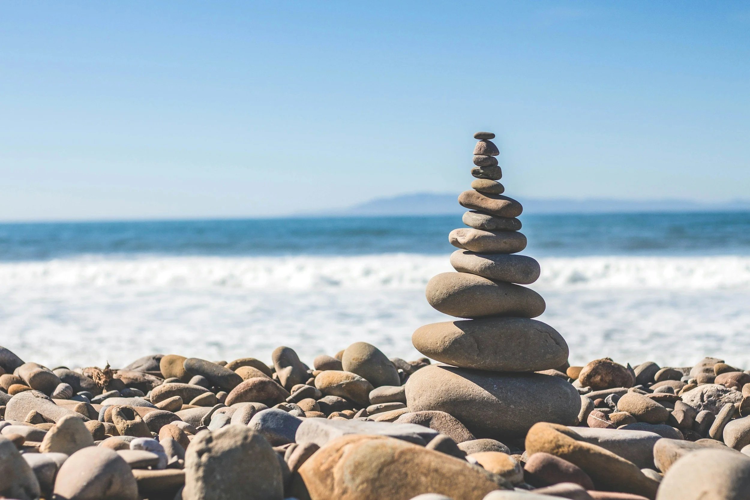 Stacked rocks on a pebbled beach with the ocean and a distant island in the background.