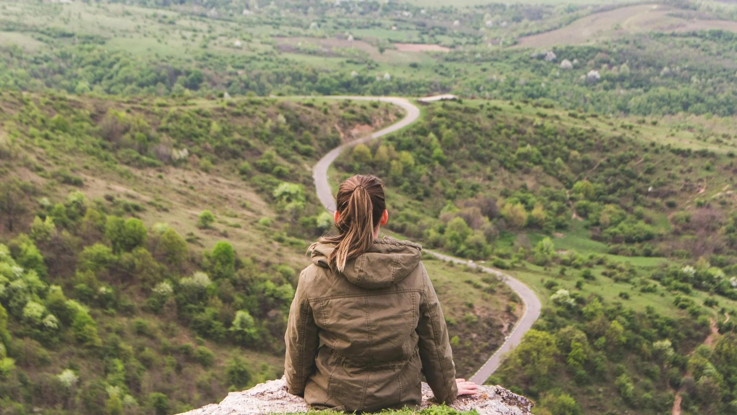 A woman with brown hair tied in a ponytail, wearing a brown jacket, sitting on a rock and facing away from the camera, looking at a winding trail through a lush green hilly landscape.