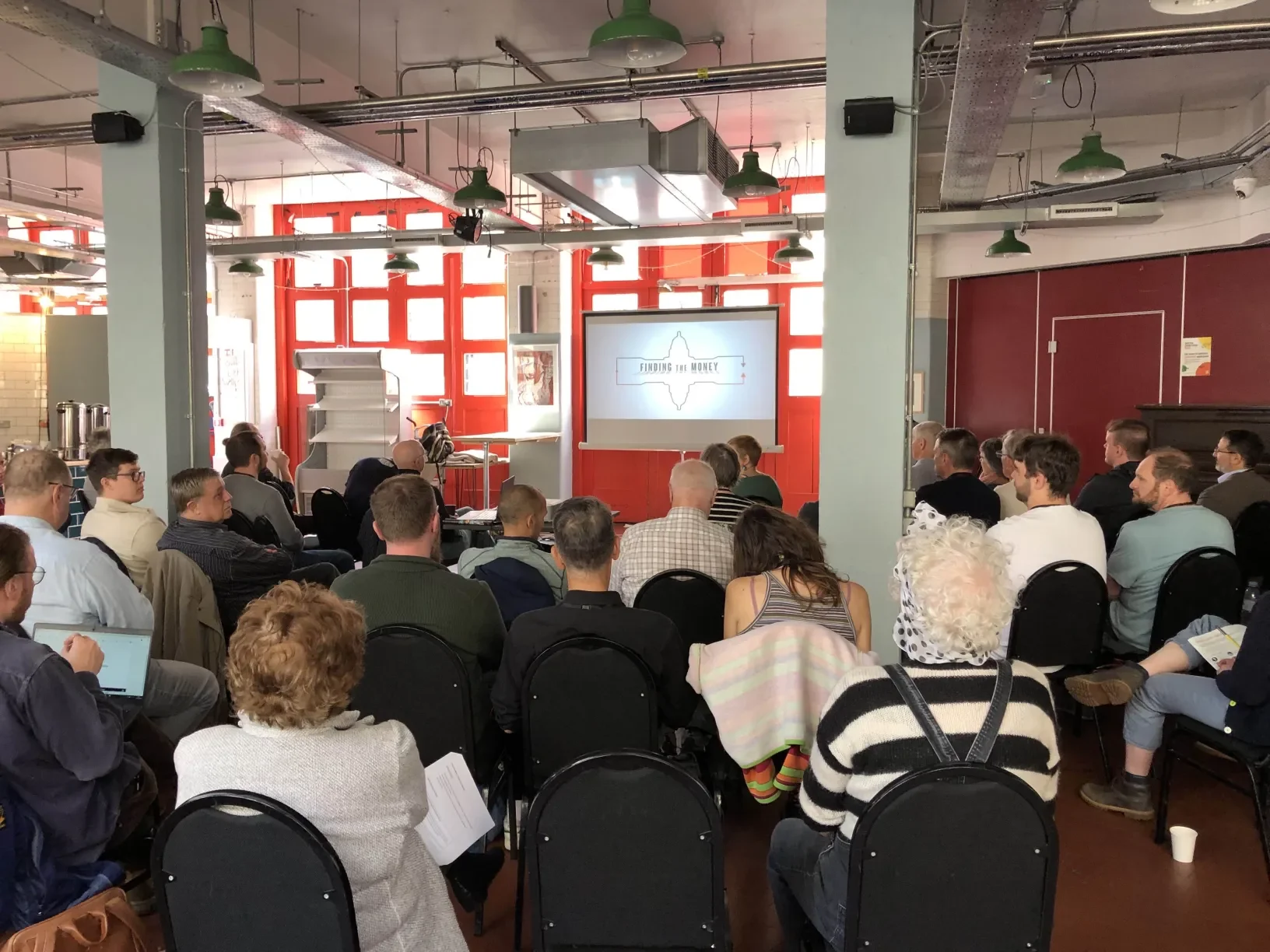 An audience of about 20 people watching a film projected onto a screen in an event space.