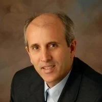 Headshot of a middle-aged man with light skin, short receding hair, wearing a dark suit jacket, white shirt, posing against a neutral background.