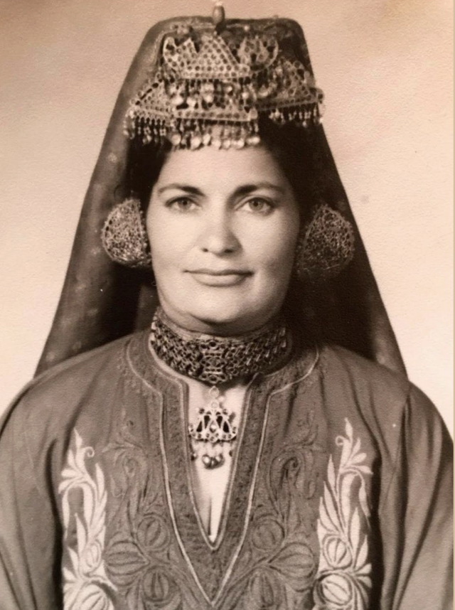 A woman wearing traditional Middle Eastern or North African attire with a decorative headpiece, large earrings, and embroidered clothing.