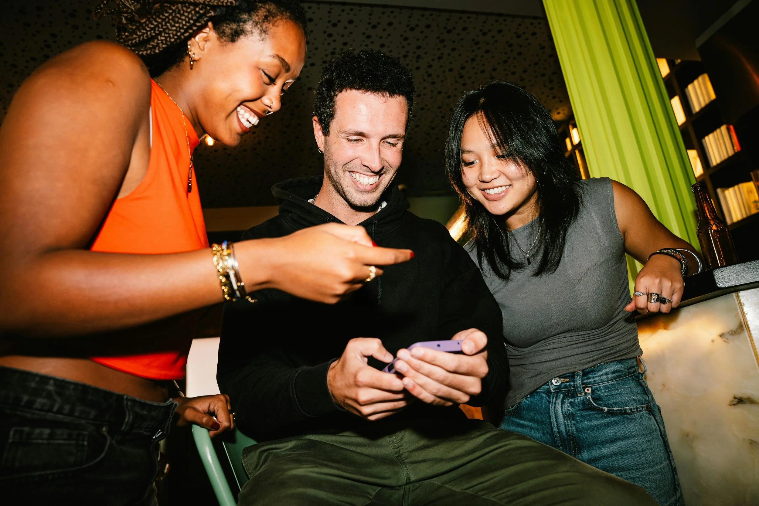 Three friends gathered around a smartphone in a lively indoor setting, smiling and looking at the phone's screen.