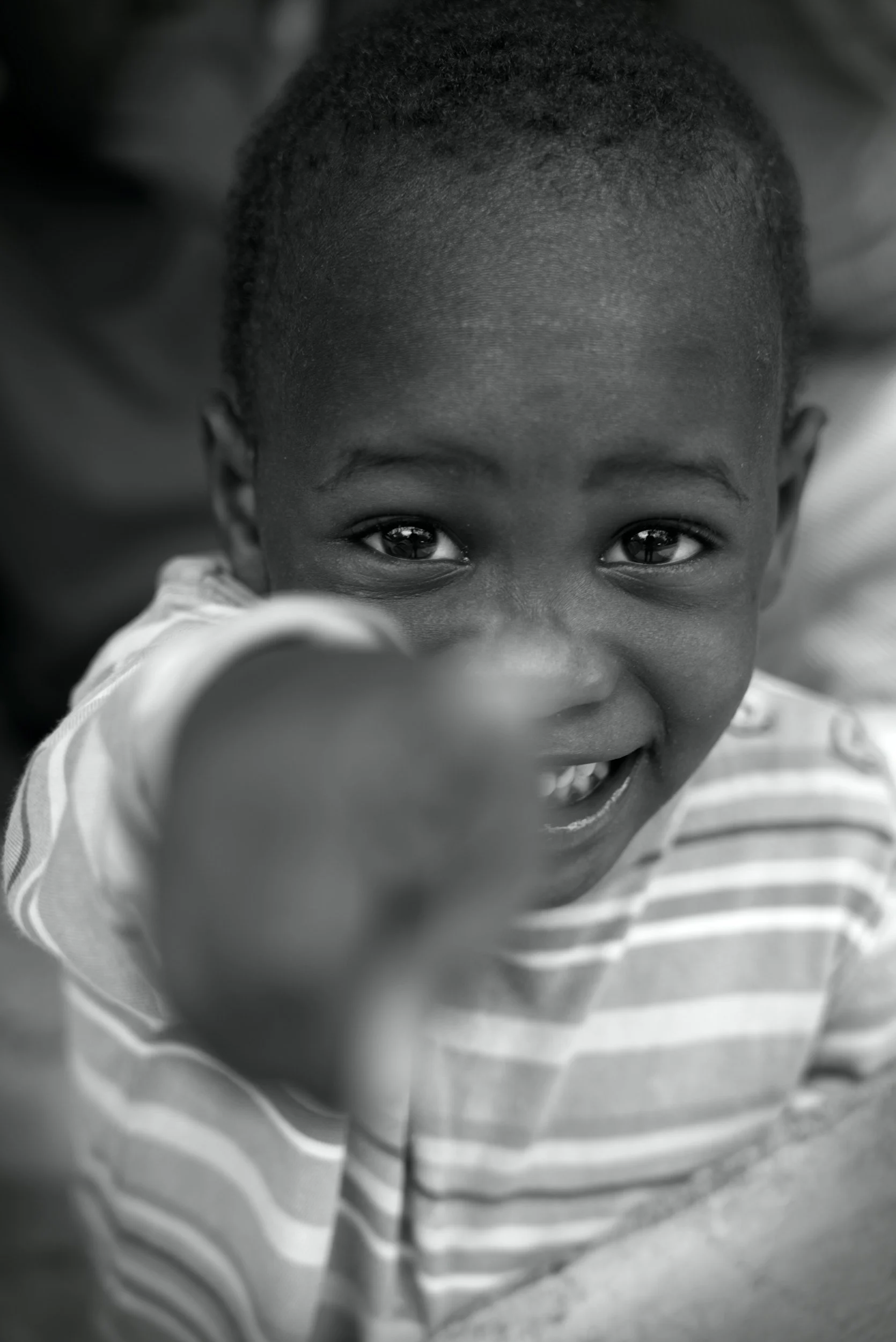 Close-up black and white photo of a smiling young African American boy with short hair, wearing a striped shirt, reaching towards the camera.