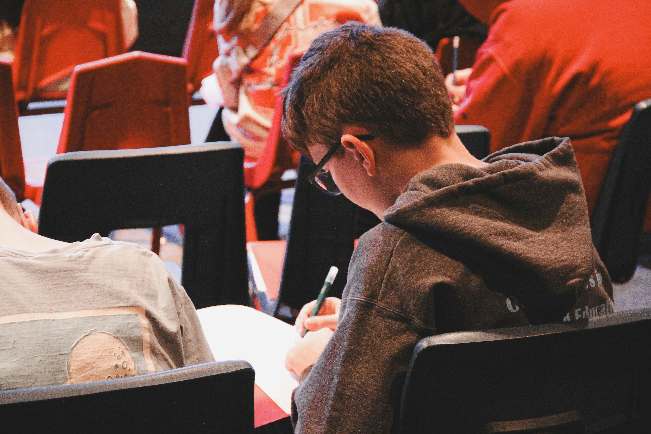 A young man with glasses and a hoodie sitting in a classroom taking notes, surrounded by other students.
