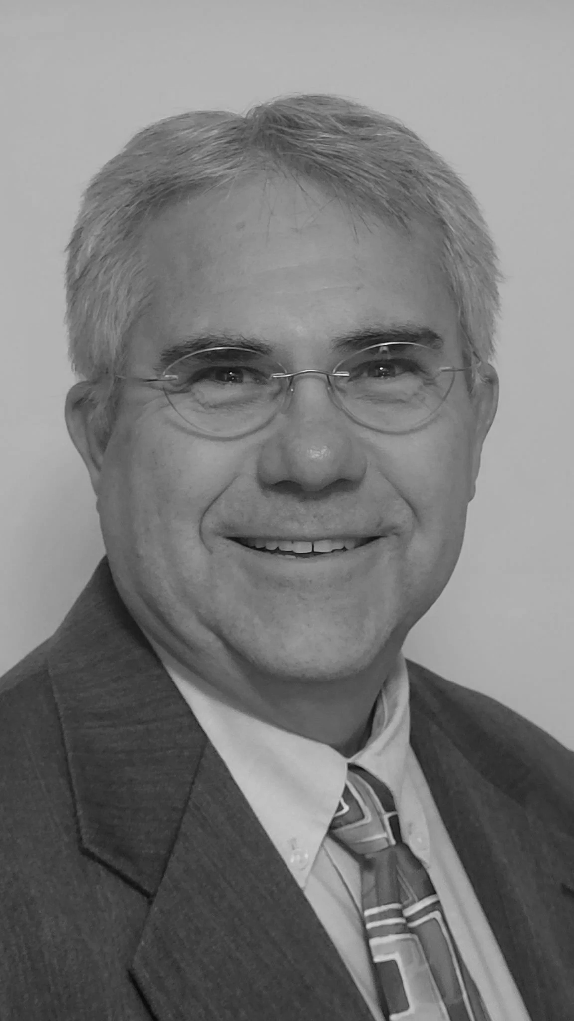 A black and white portrait of a smiling man in glasses wearing a suit and patterned tie.