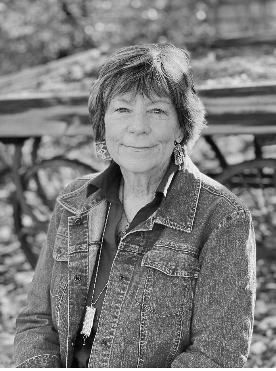 An elderly woman with short hair, smiling, wearing a denim jacket and earrings, sitting outdoors with a blurred background.