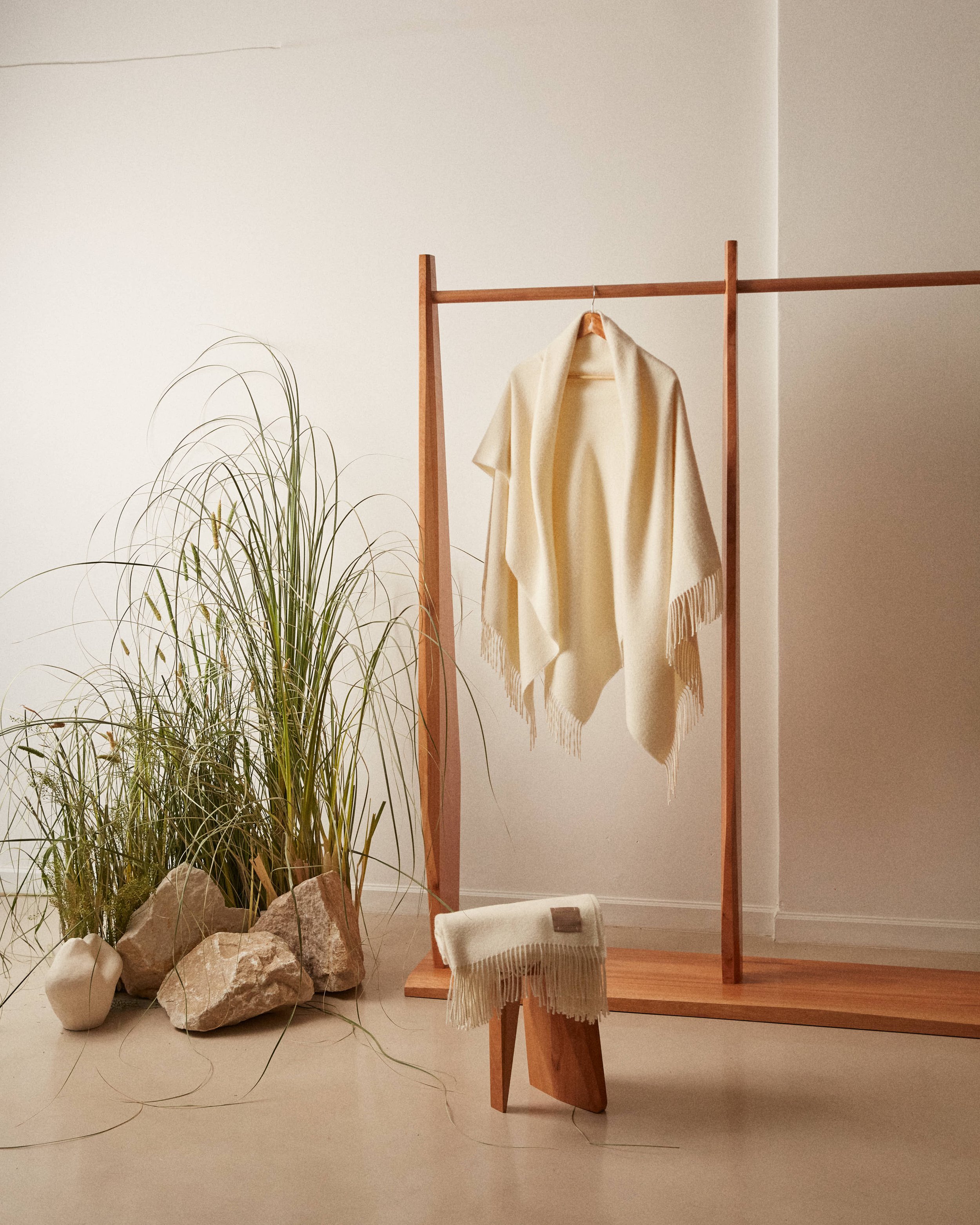 A minimalist clothing display with a wooden rack holding a cream-colored shawl, a plant with tall green leaves and rocks on the floor, and a small wooden stool with a fringed cream piece on top.