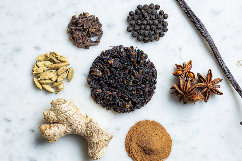 Un assortiment d'épices séchées sur une surface blanche, incluant du gingembre, de la poudre de curry, des graines de cardamome, du clou de girofle, de la cannelle, du star anise, des noix de muscade et des piments séchés.